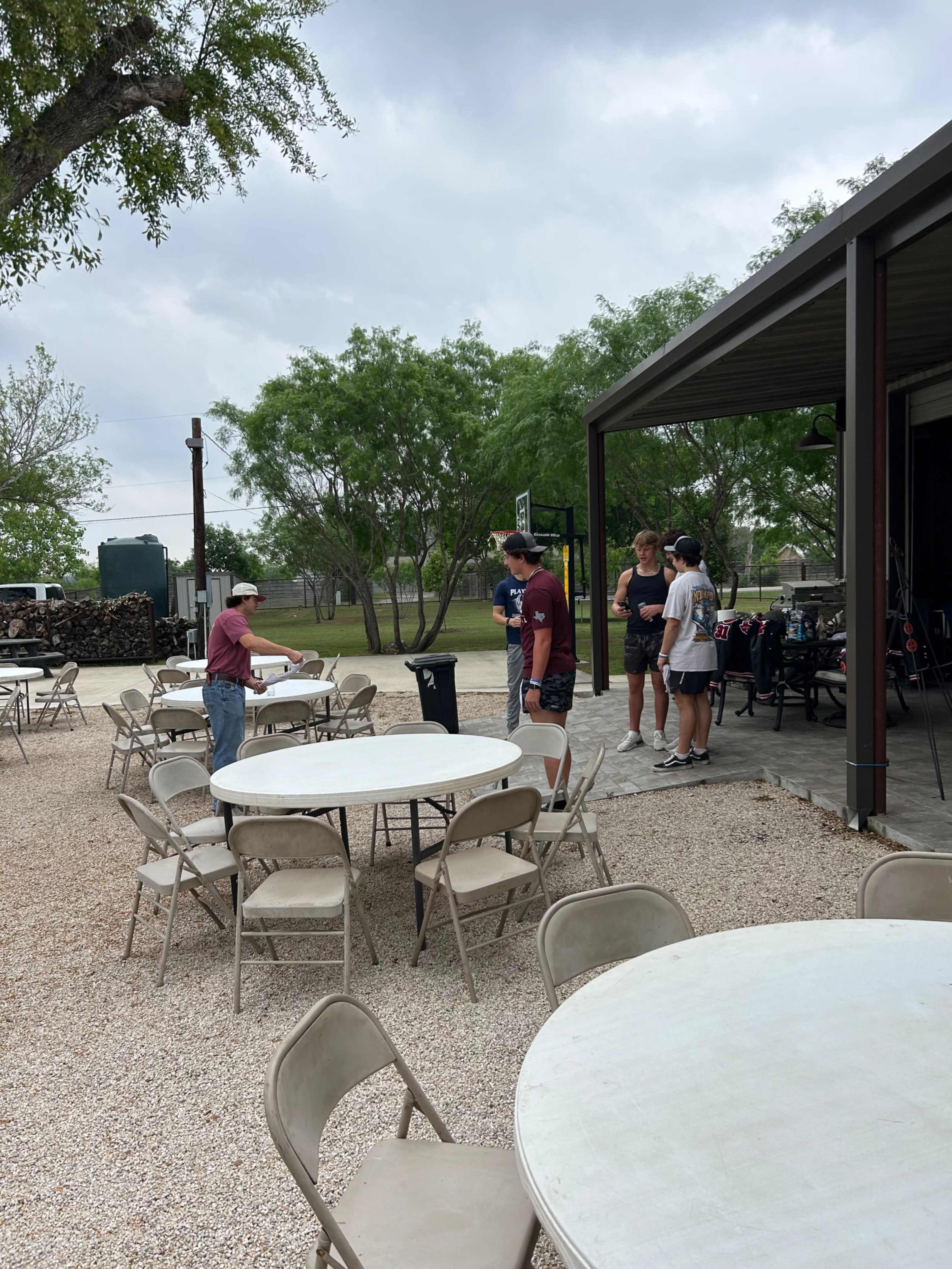 A group of five people stands among tables outside a building, while another person prepares a trash can nearby.