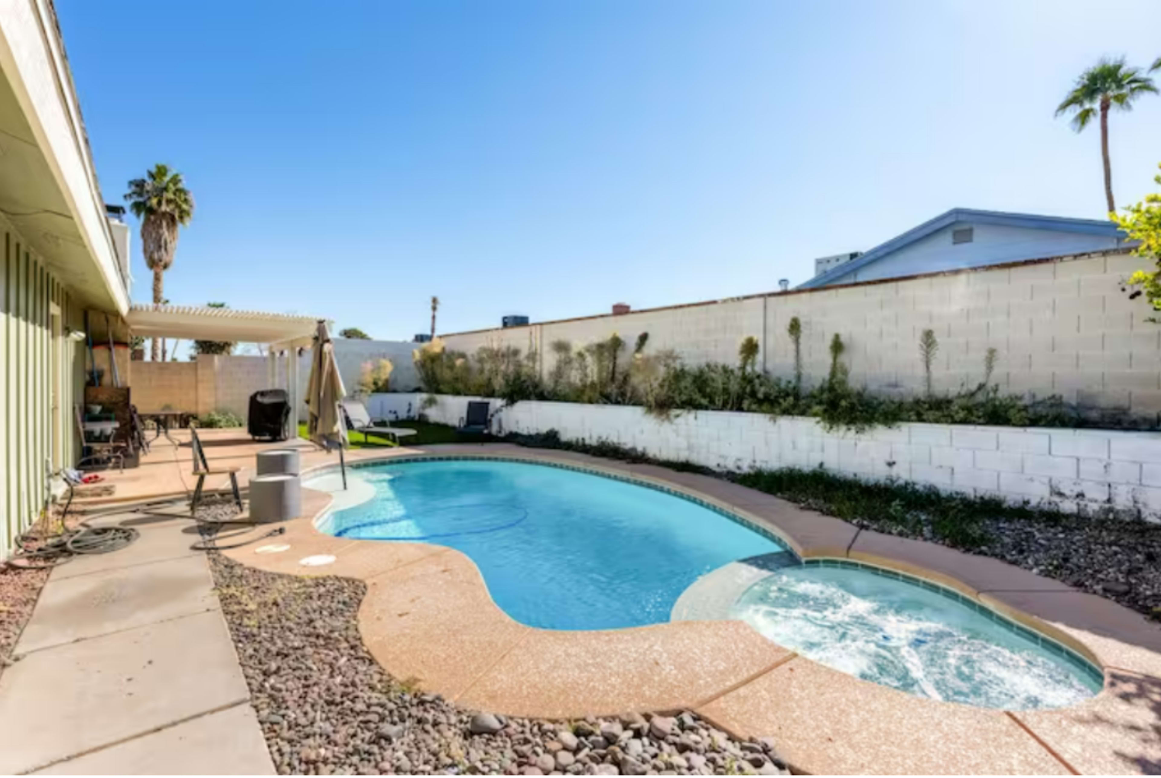 A backyard with a curved swimming pool and a hot tub, surrounded by a stone patio and landscaped areas.