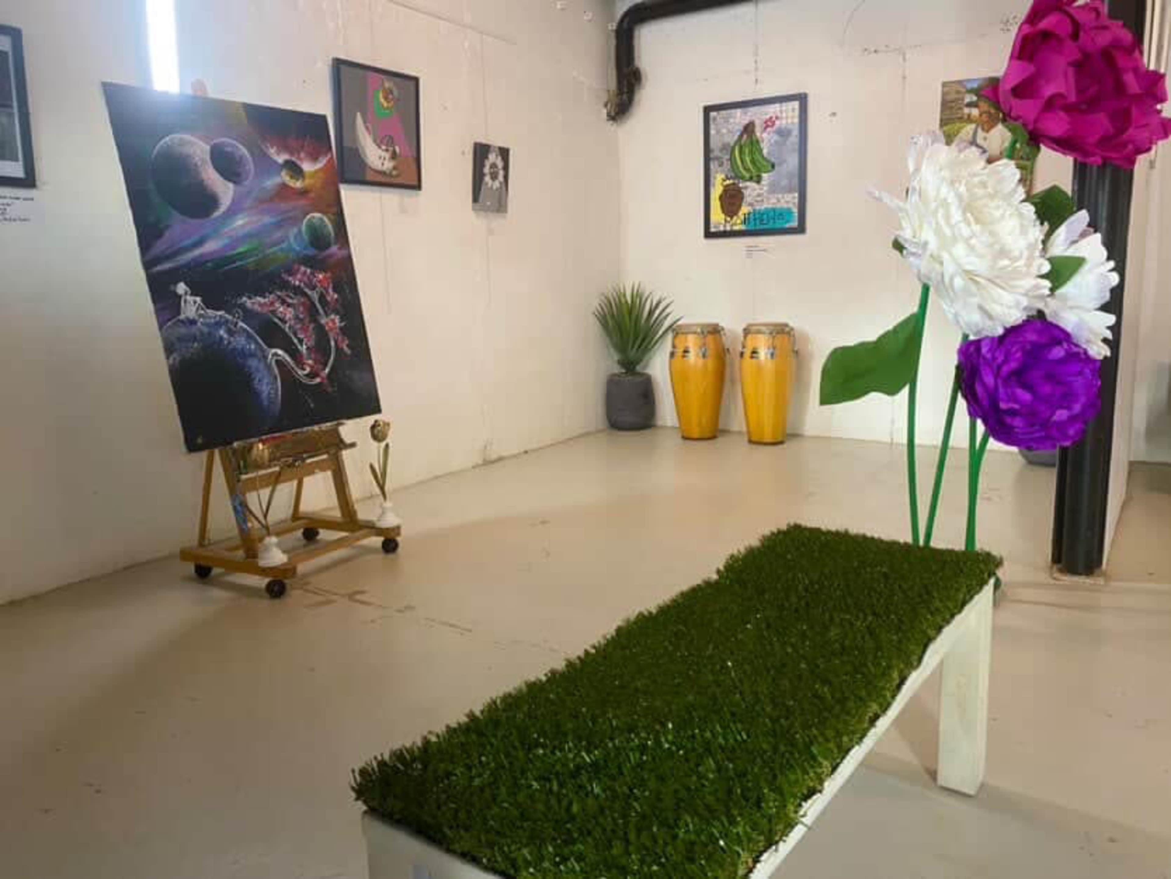 The image shows an art gallery space featuring a colorful painting on an easel, framed artworks on the walls, a green bench, and conga drums in the background.