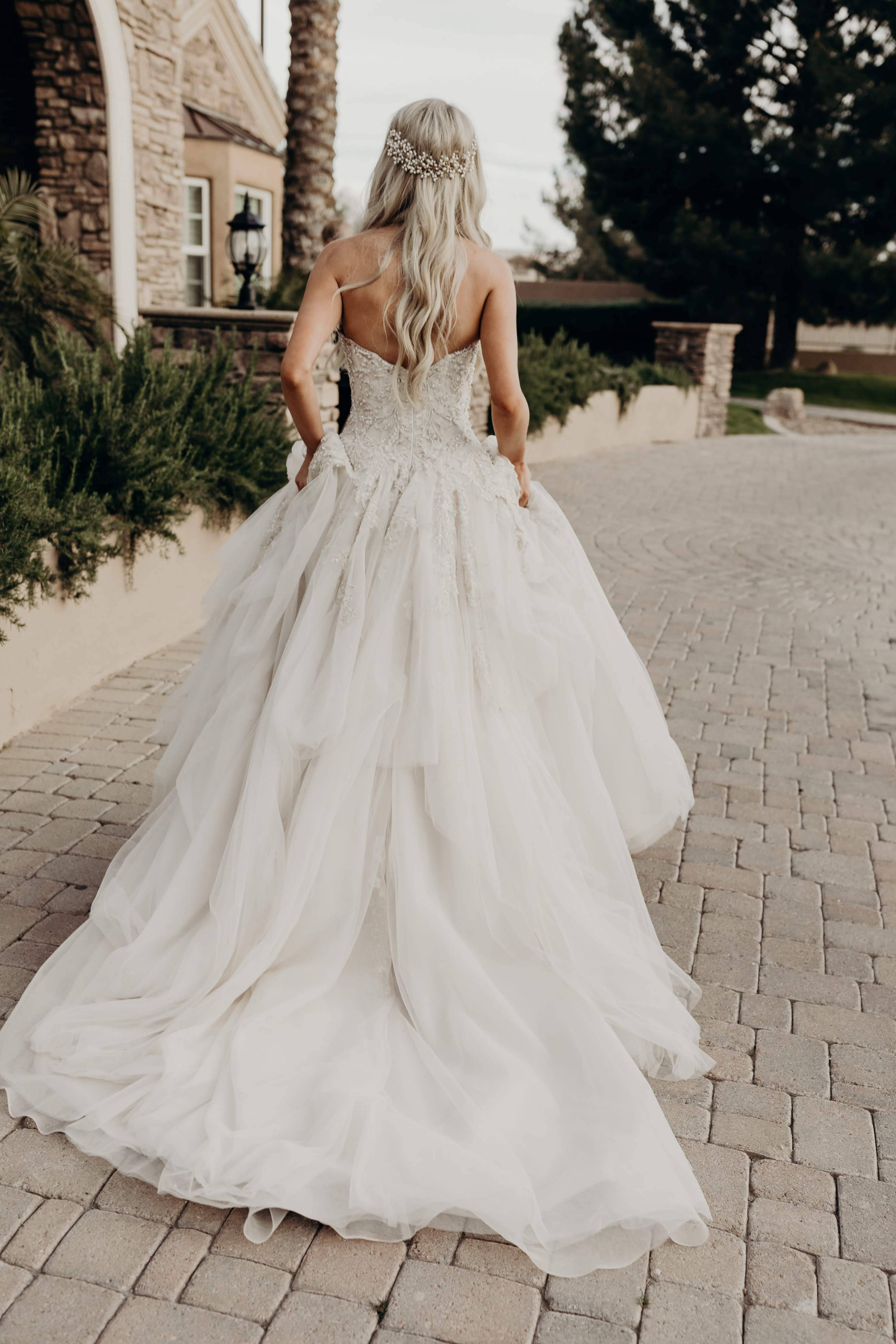 A woman with long blonde hair, wearing a flowing white wedding gown, walks away along a stone pathway lined with greenery and palm trees.