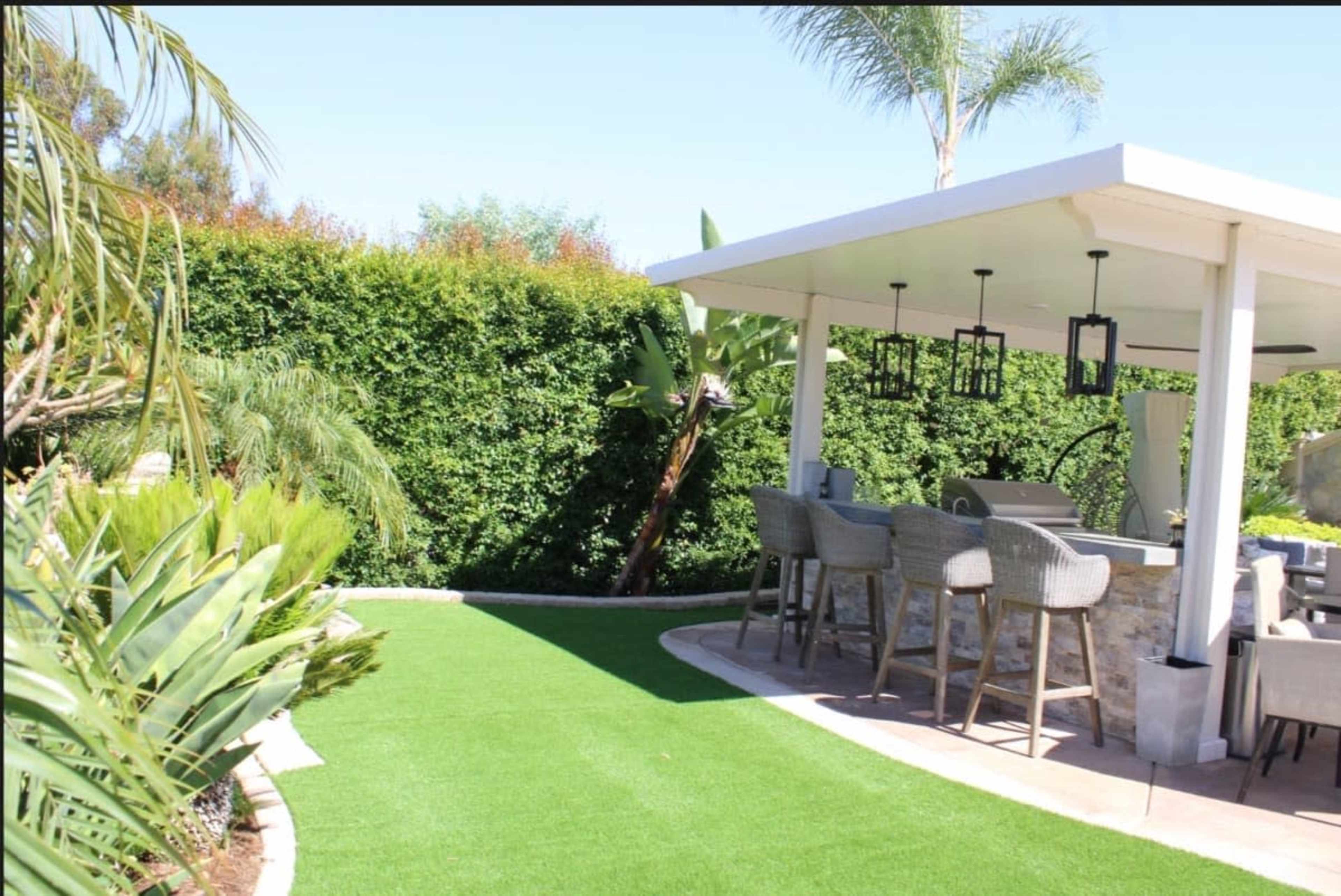 A landscaped outdoor area features a green artificial lawn, a stone bar under a white gazebo, and seating surrounded by lush greenery.