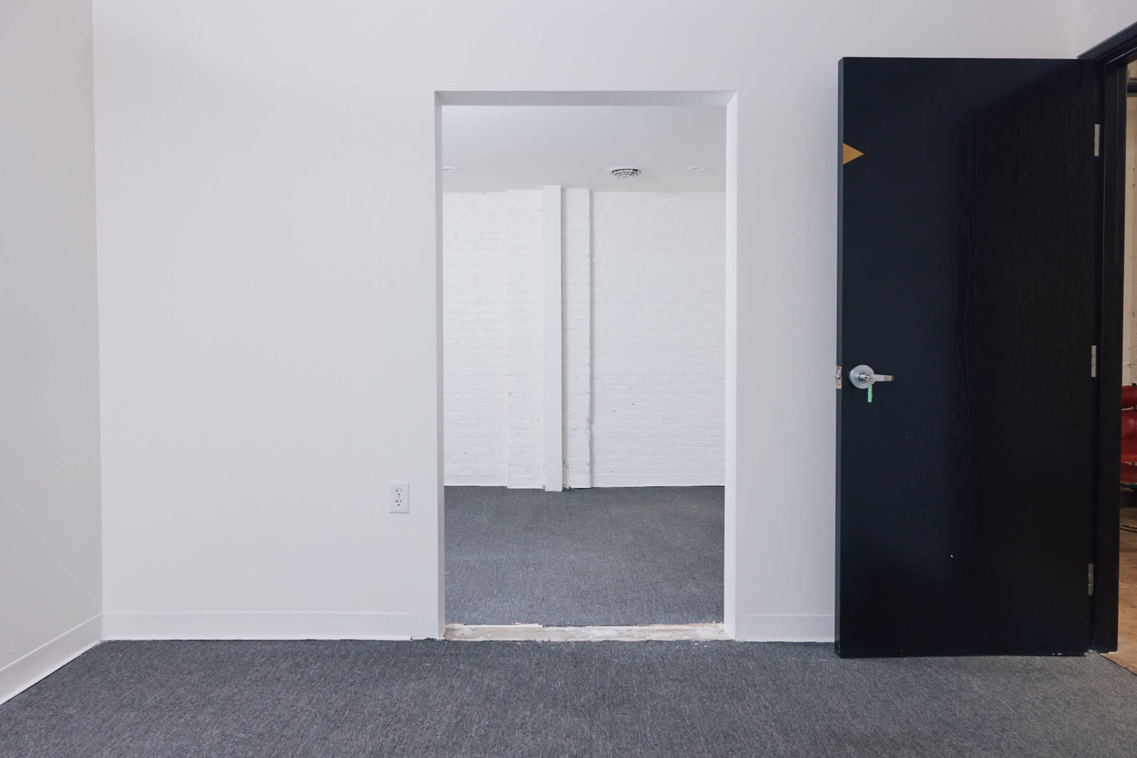 The image shows a white room with a doorway leading to another room, featuring carpeted flooring and a black door on the right side.