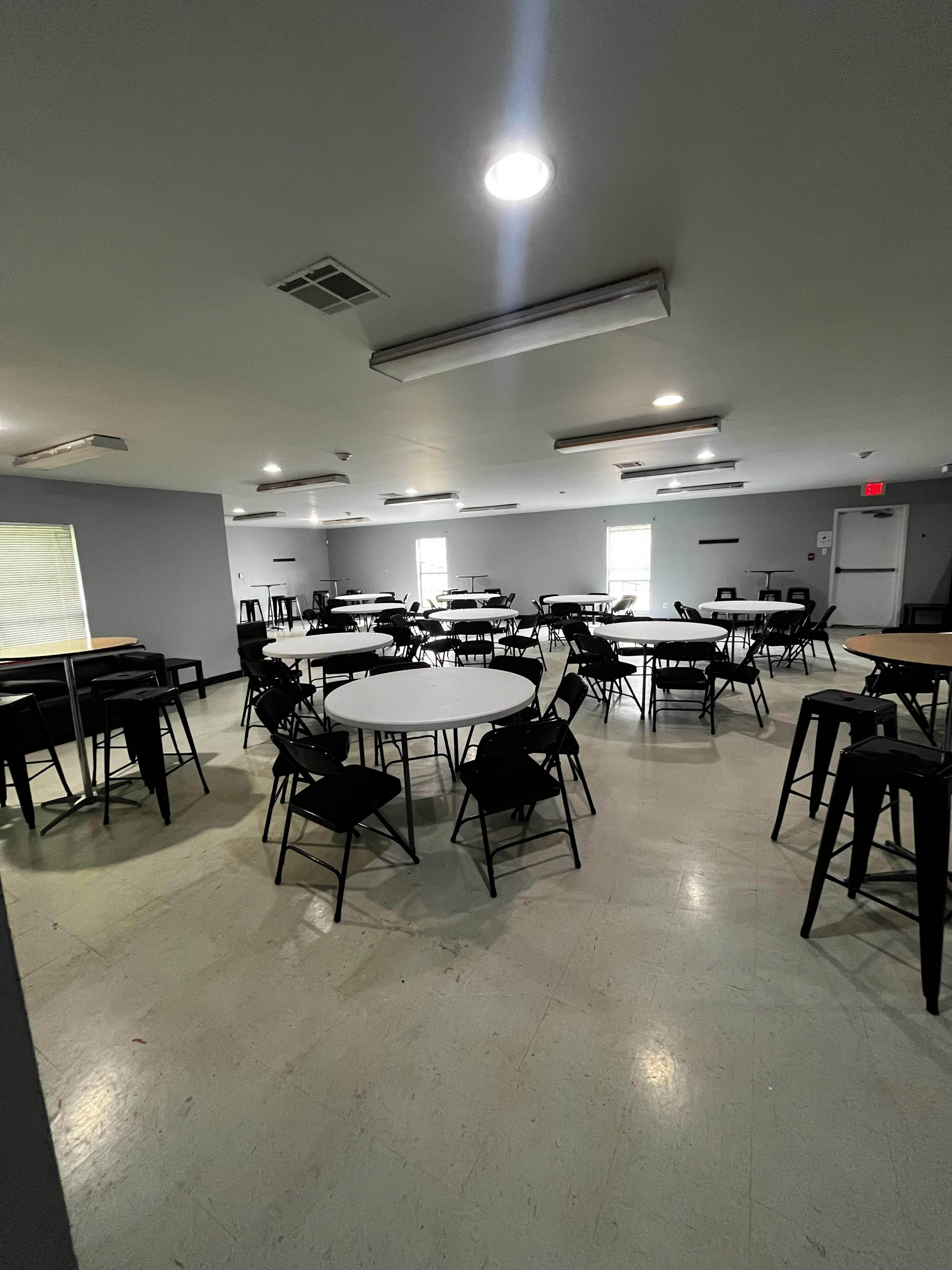 A spacious, well-lit room contains several round tables with black chairs arranged throughout.