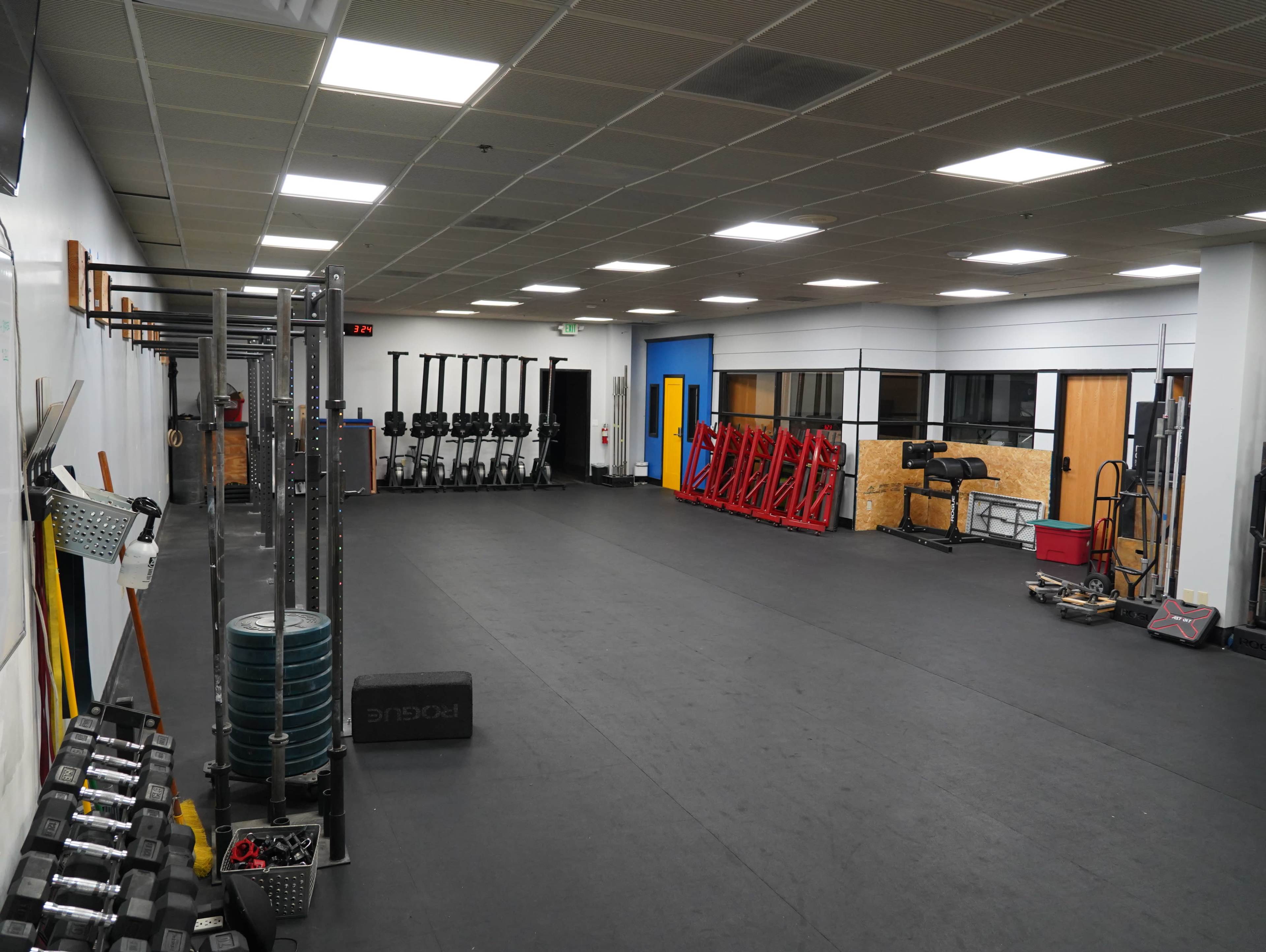 The image shows a spacious gym with various fitness equipment, including dumbbells on one side and rowing machines lined up against the wall.