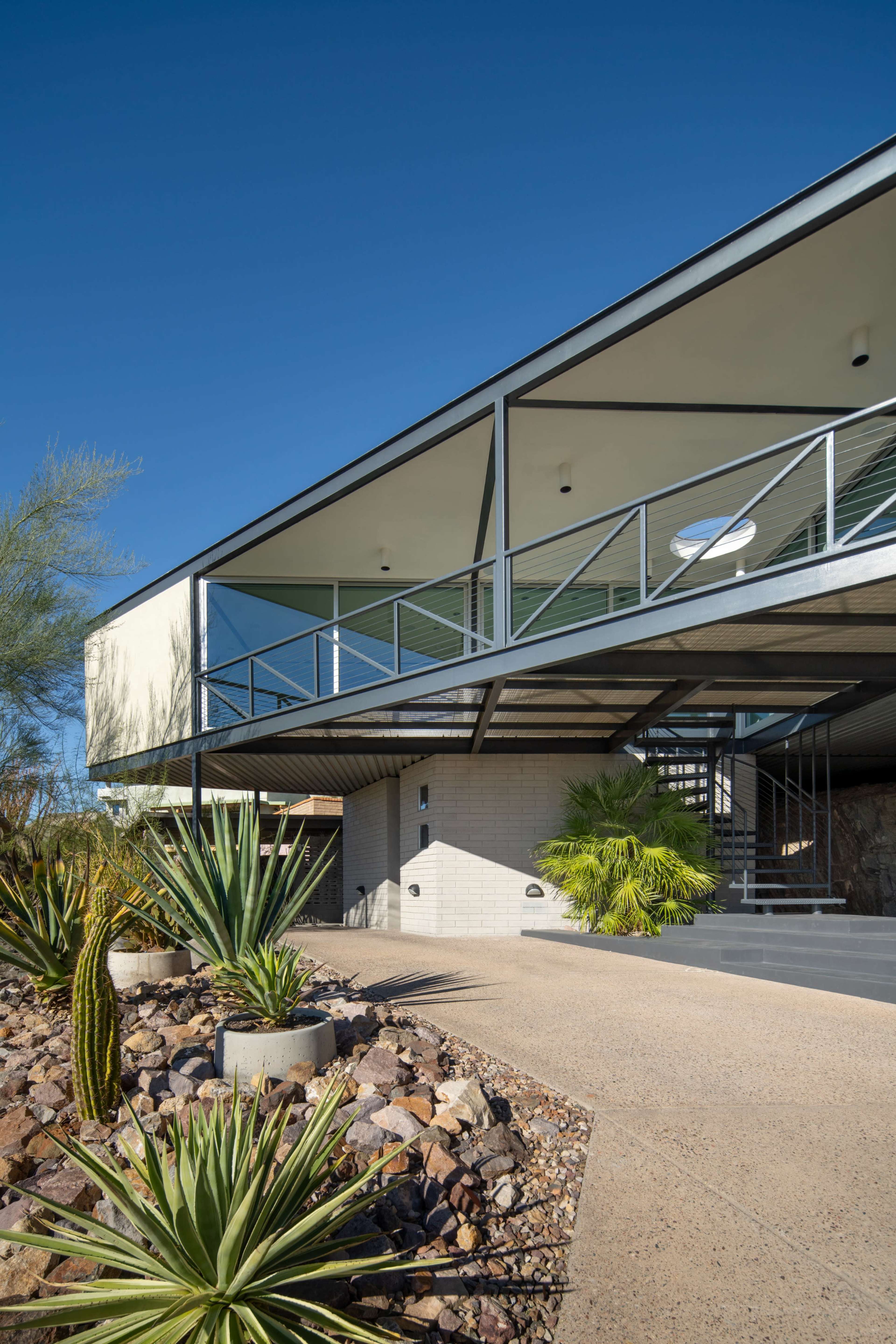 A modern house with a cantilevered upper level features a landscaped entrance with various desert plants and rocks.