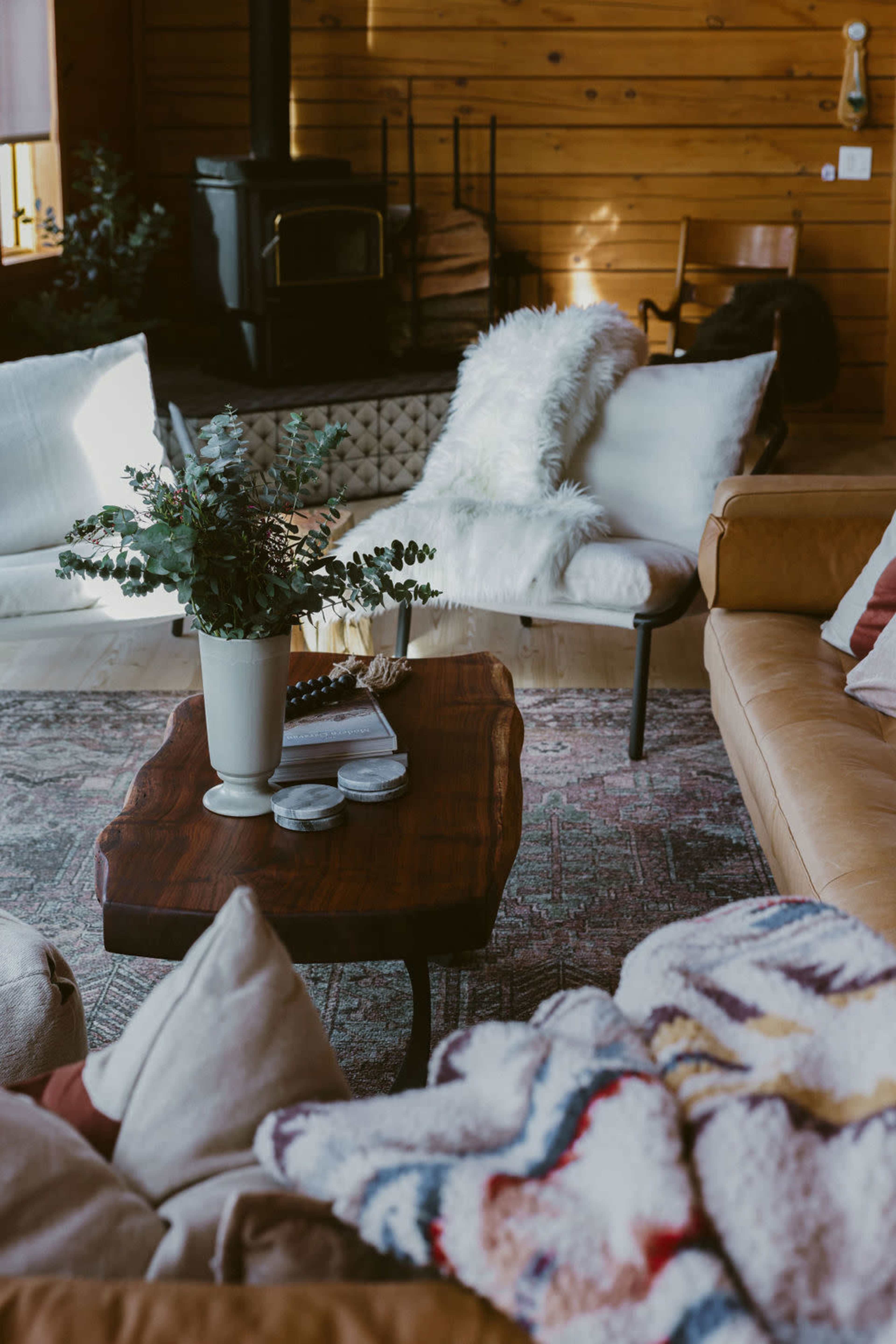 A wooden coffee table topped with a white vase and candles sits in a cozy living room with a mix of upholstered furniture and wooden walls.