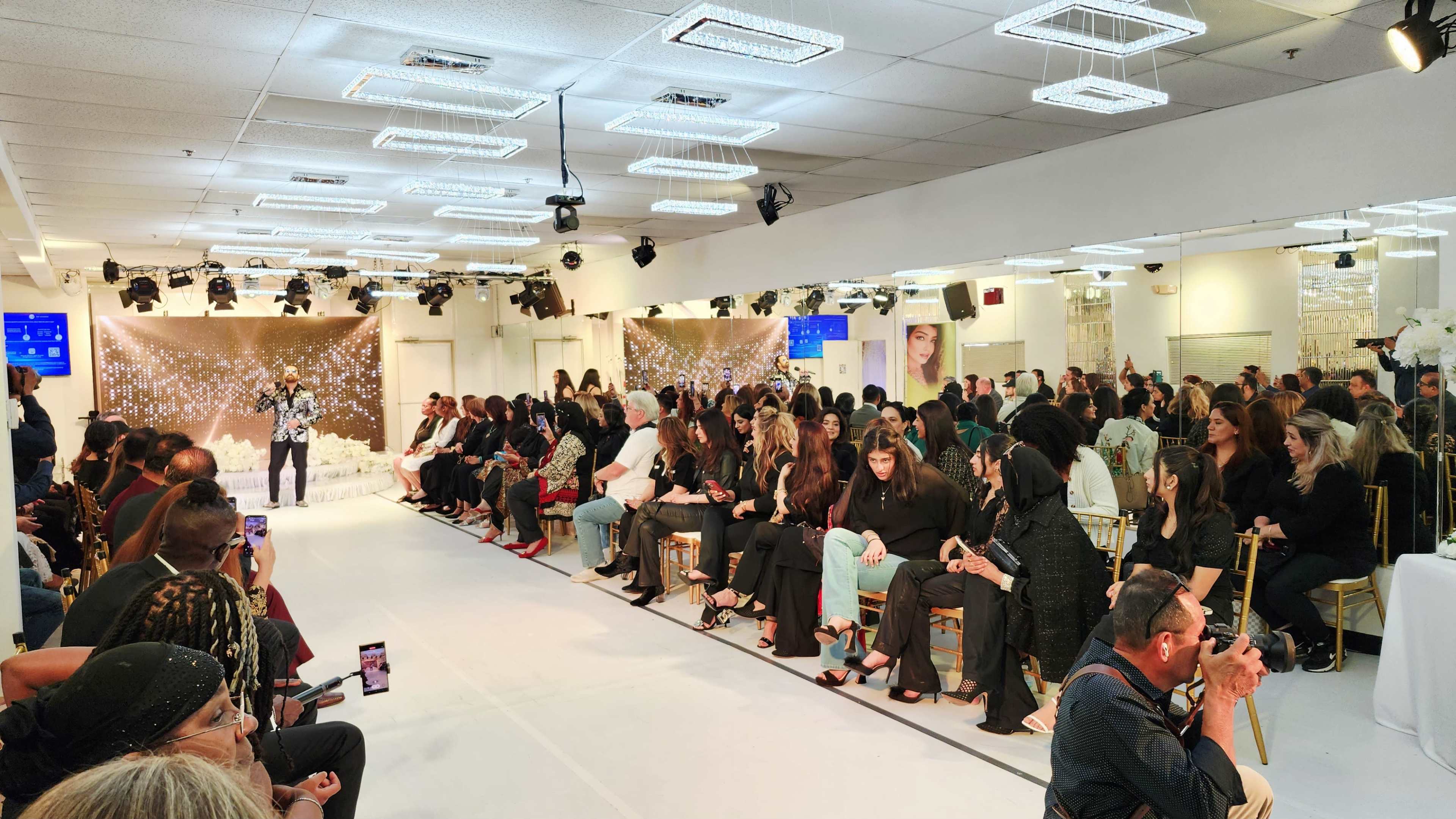 A crowded fashion show venue features attendees seated in rows, with a model preparing to walk the runway in front of a decorative backdrop.