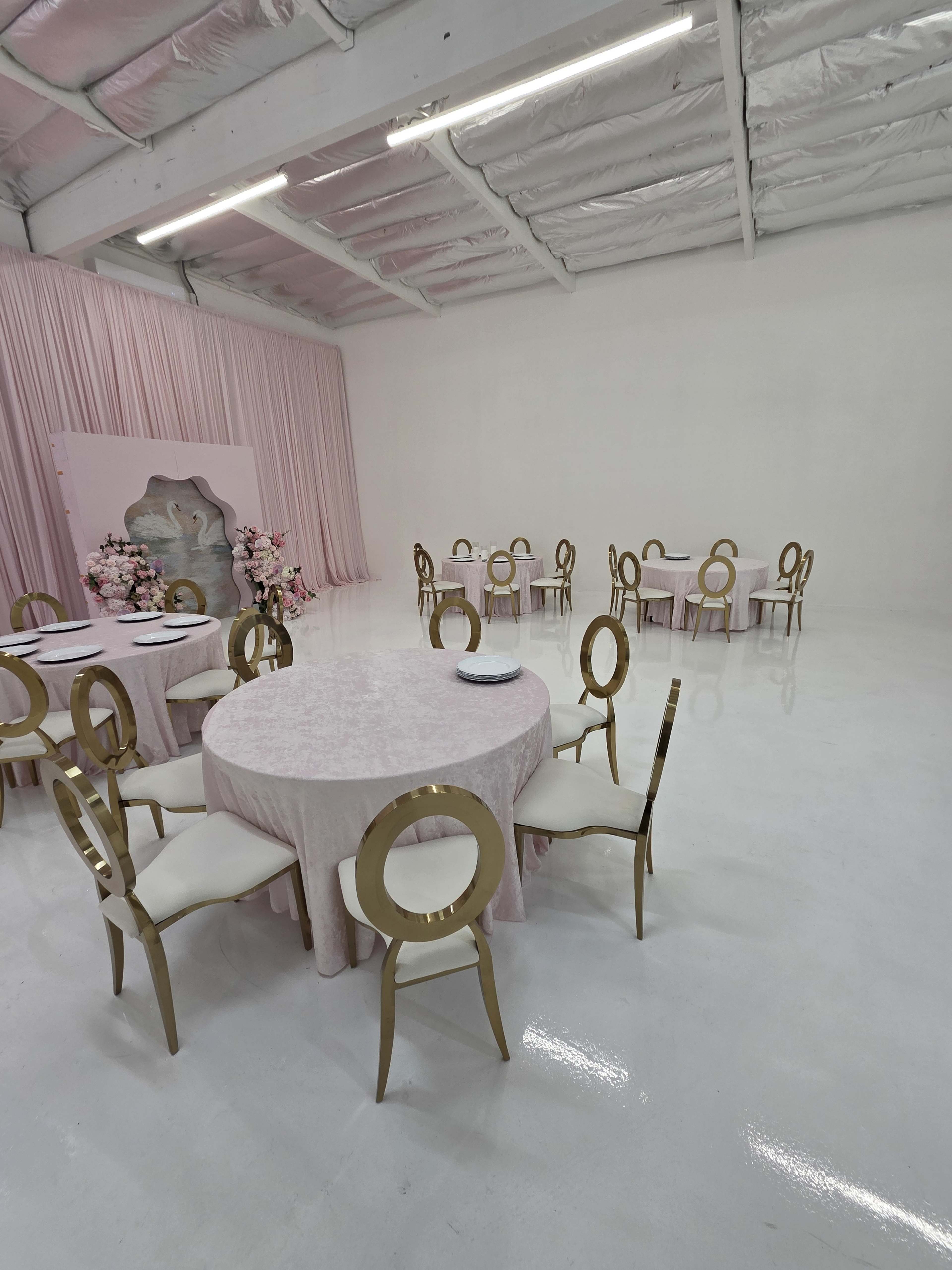 The image shows a spacious venue with white walls and a shiny floor, featuring multiple round tables draped in light pink tablecloths and gold chairs arranged neatly.