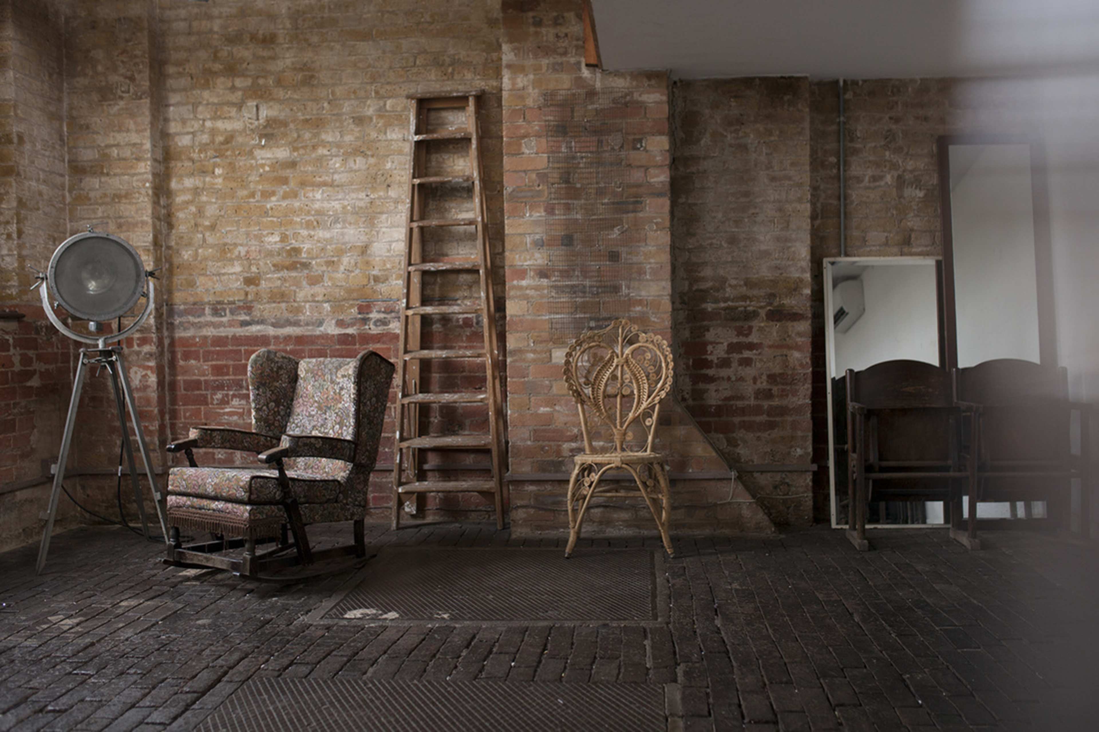 A minimalist interior space with two armchairs, a wooden ladder, a large light fixture, and a mirror against a brick wall.