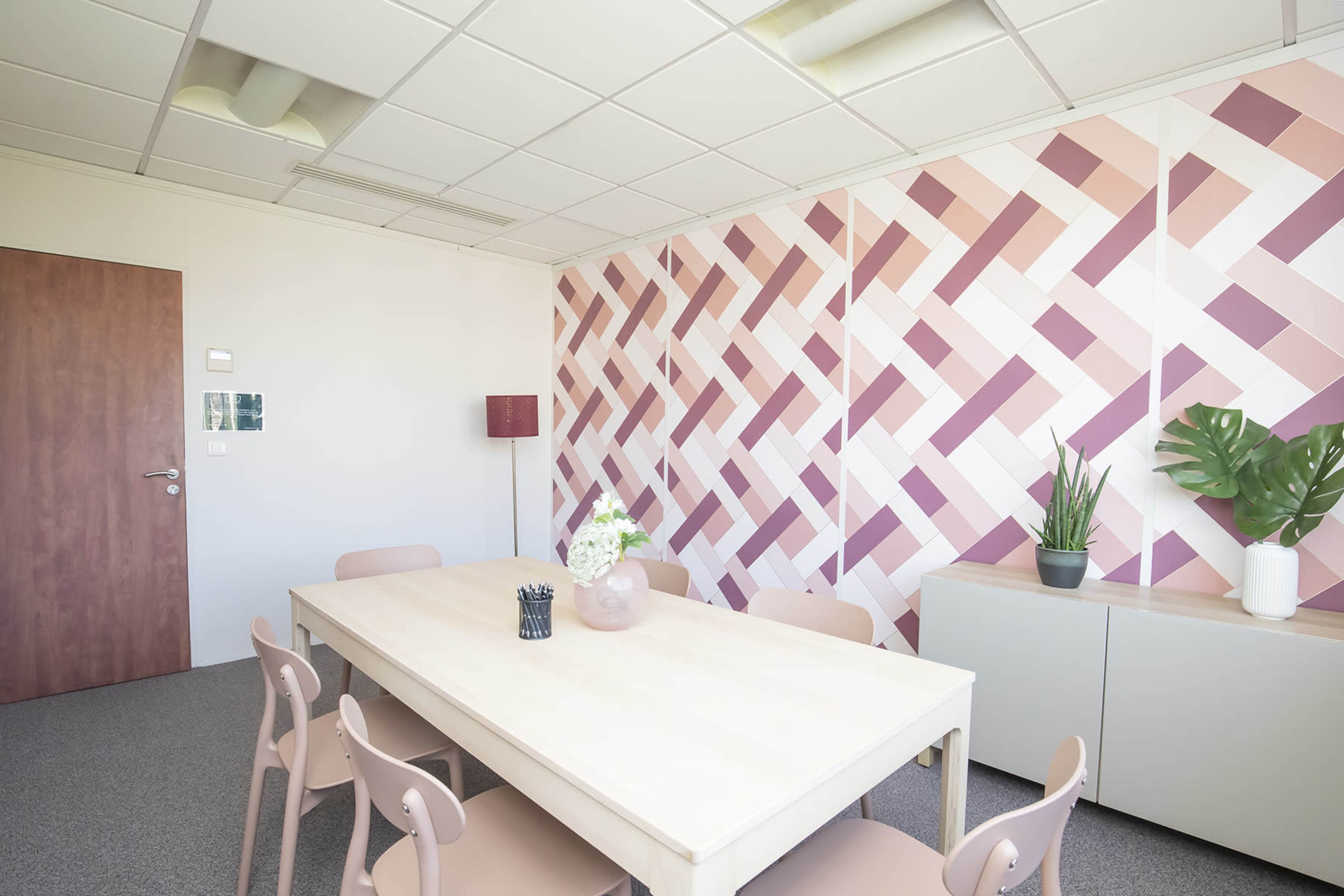 A conference room features a wooden table surrounded by light-colored chairs, with a patterned pink and white wall, a potted plant, and a floor lamp.
