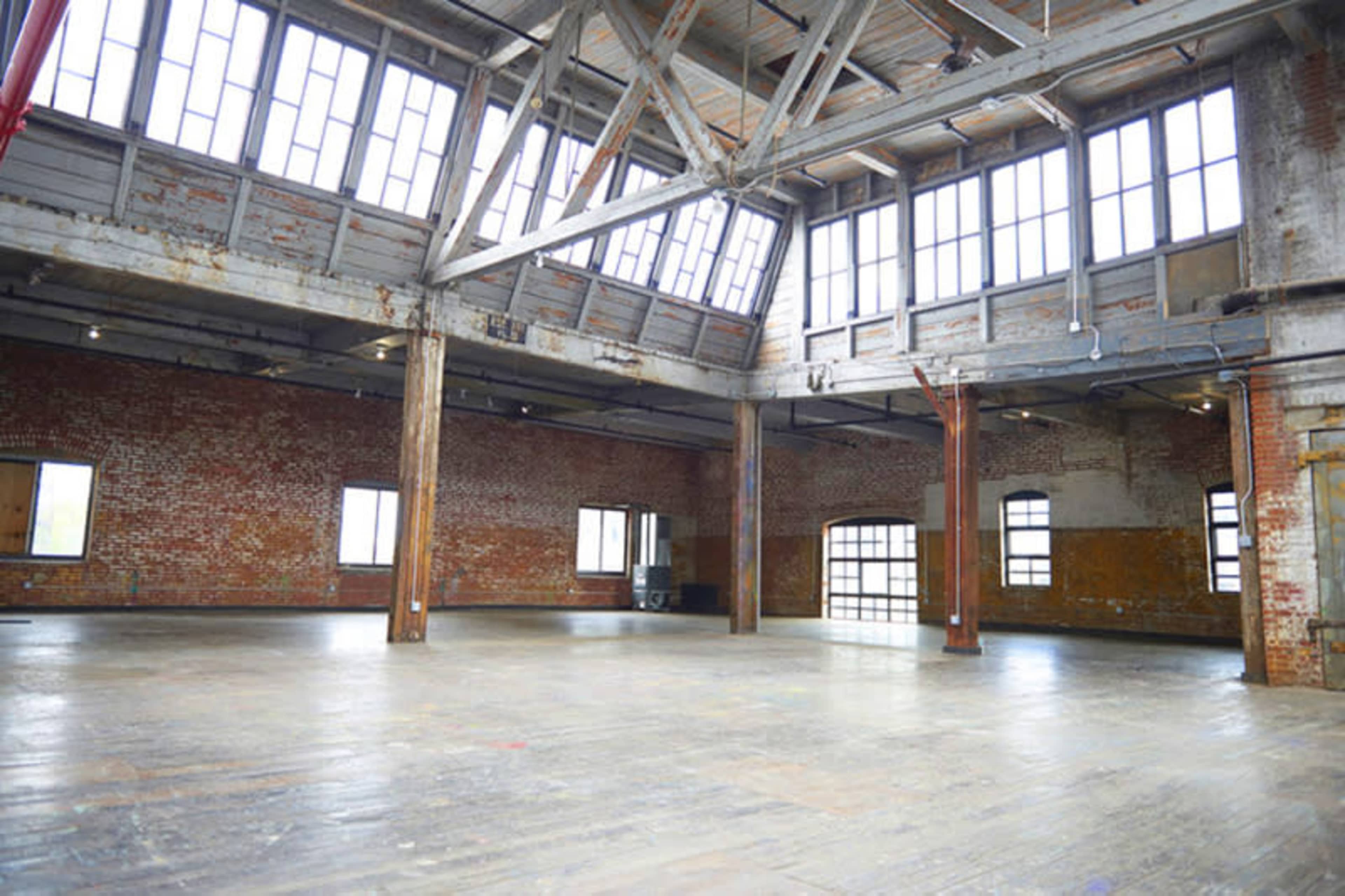 The image shows a large, open space with exposed brick walls, wooden beams, and large windows allowing natural light to flood the interior.