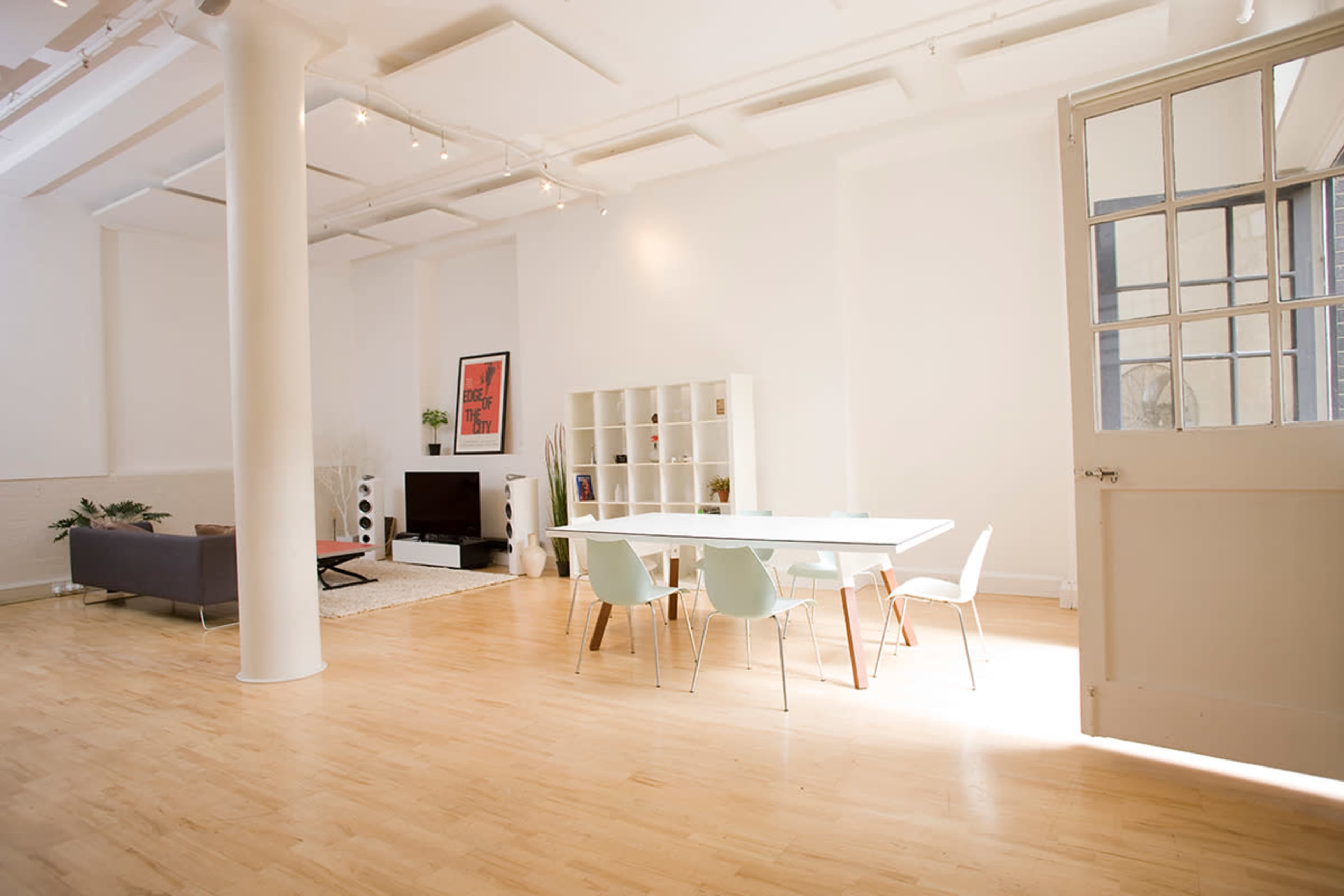 A bright, open-concept living space featuring a dining area with a white table surrounded by chairs, a gray sofa, a television, and a large window.