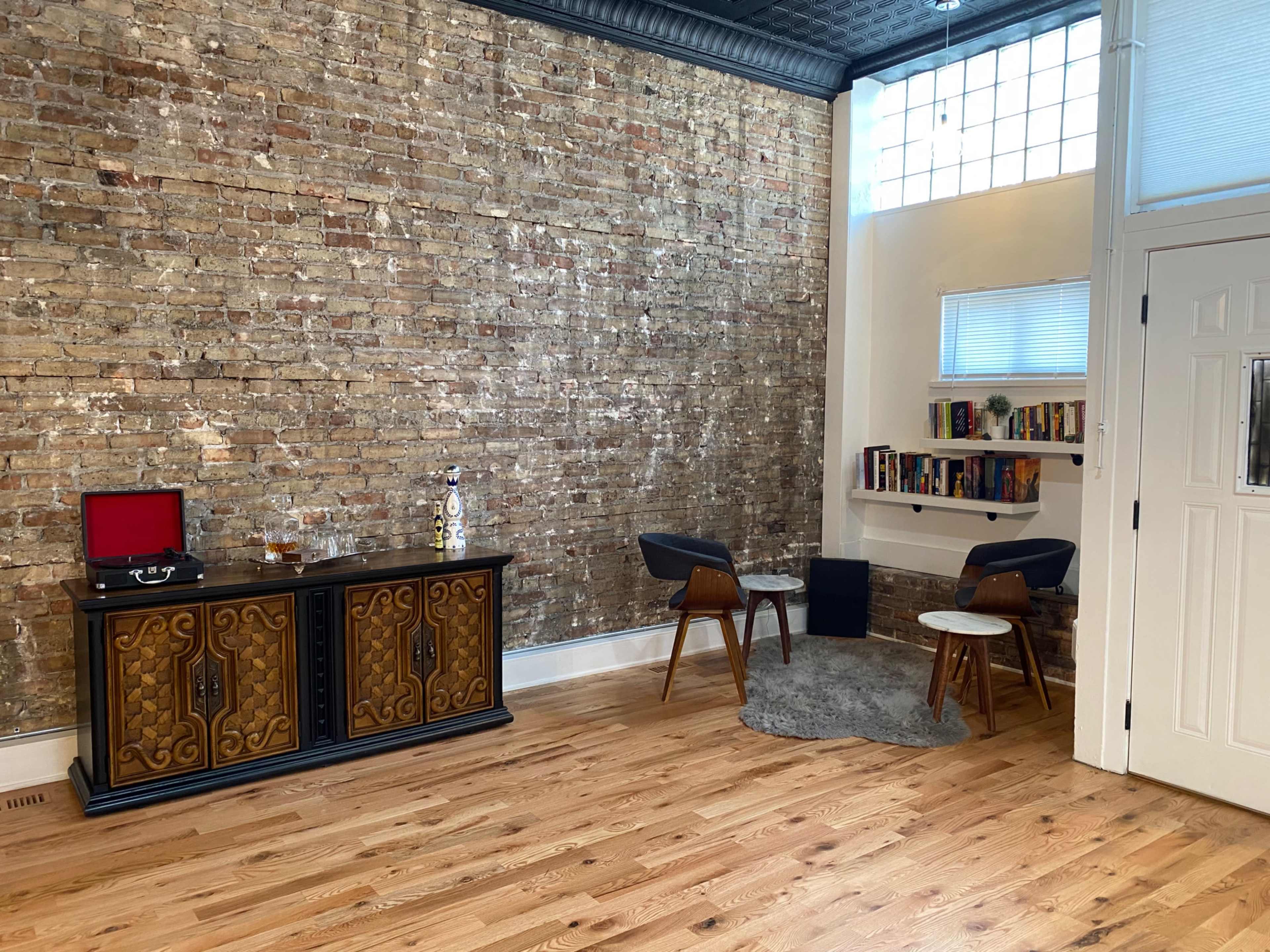 The room features a wooden floor with a brick wall, a vintage cabinet, a record player, two chairs, and a bookshelf filled with books.