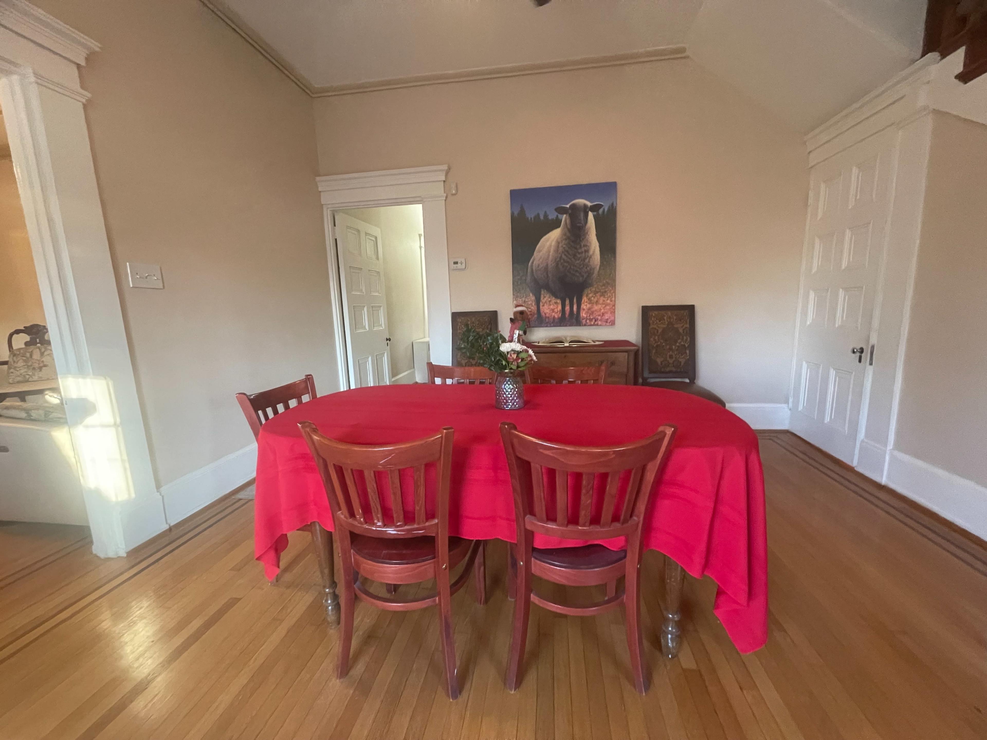 A round table covered with a red tablecloth is set in a room with wood floors and a painting of a sheep on the wall.