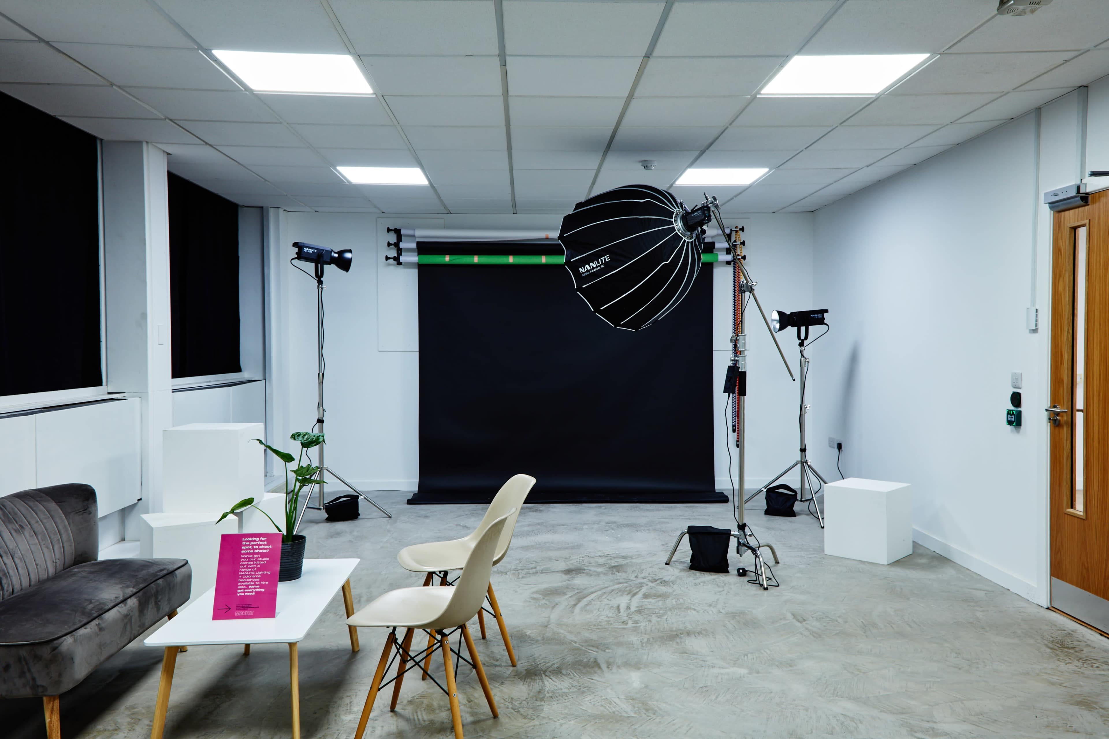The image shows a photography studio with a black backdrop, lighting equipment, a couch, a chair, and a small table.