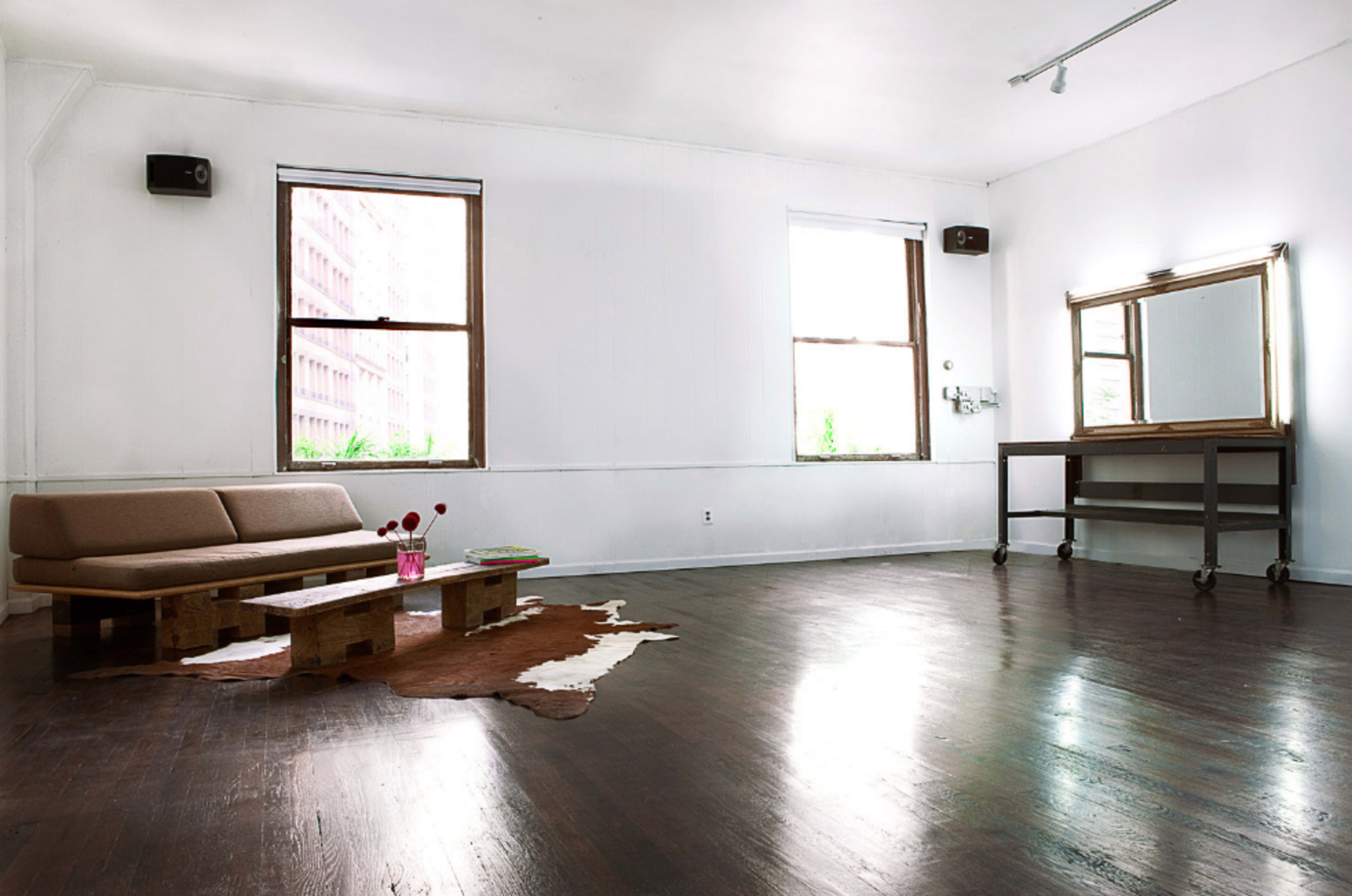 A spacious, minimalist room with large windows, a low wooden sofa, a small table with flowers, and a mirror on a rolling stand.