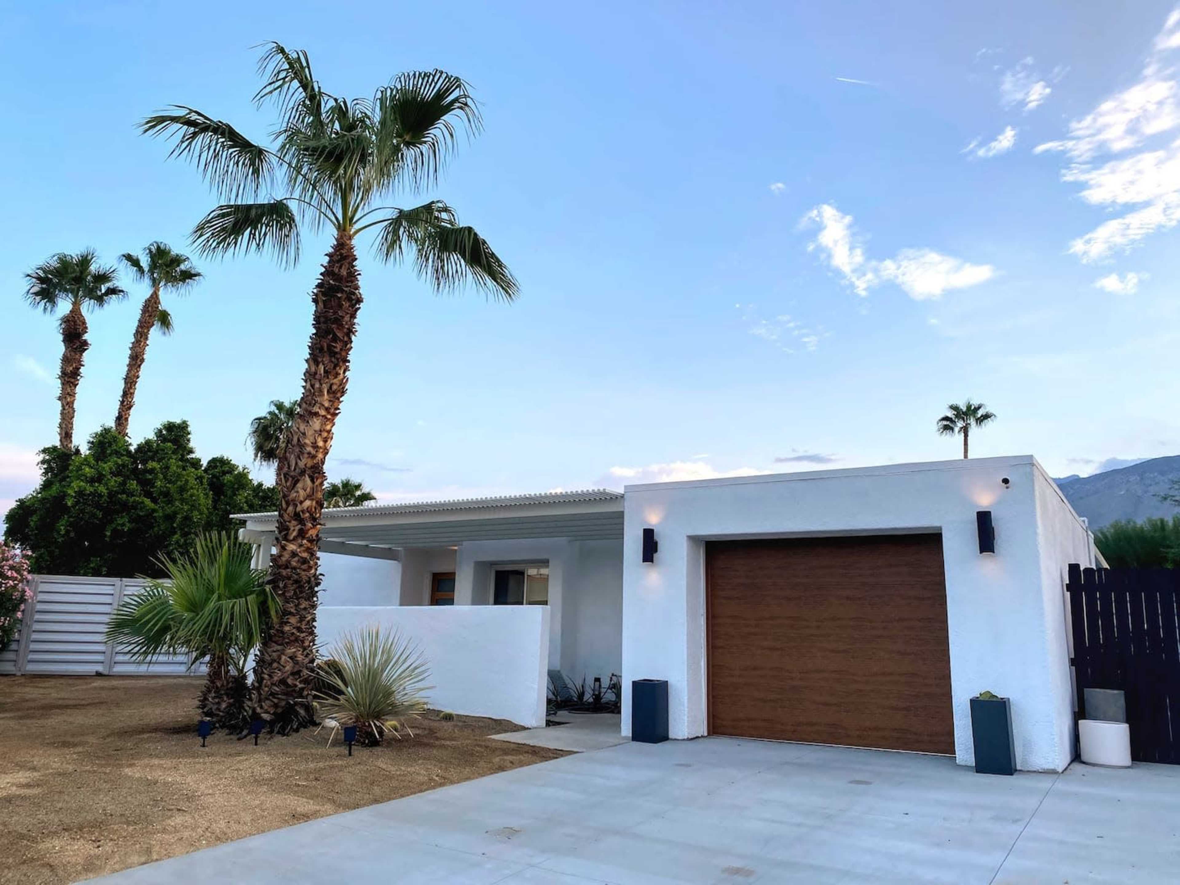 A modern white house with a wooden garage door is surrounded by palm trees and a landscaped yard.