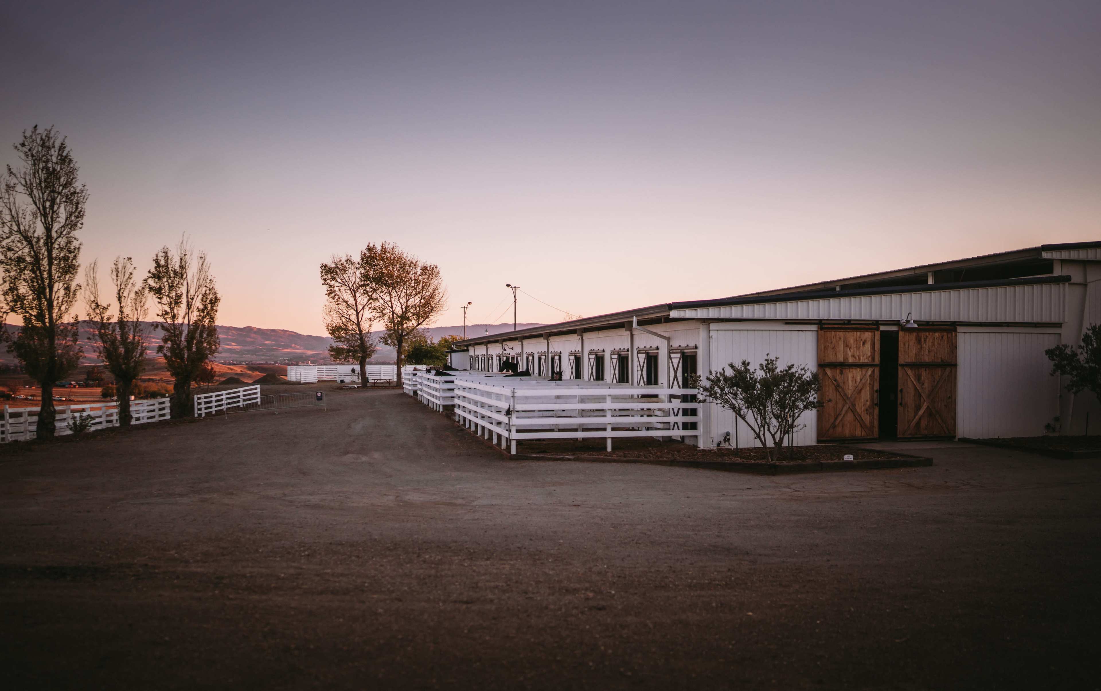 A gravel path leads to a white barn with wooden doors, surrounded by trees against a sunset sky.