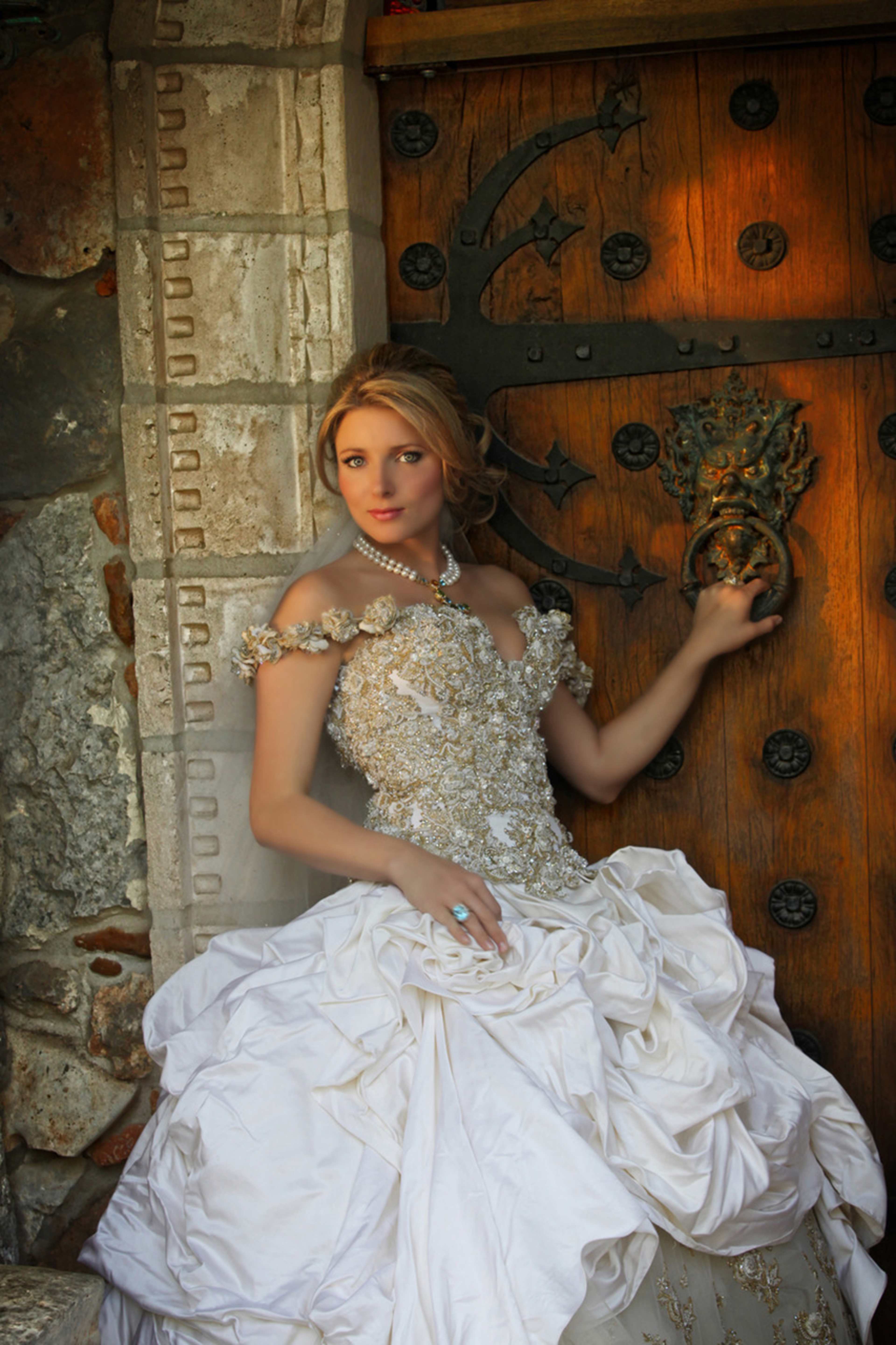 A woman in a detailed wedding gown poses beside an ornate wooden door.
