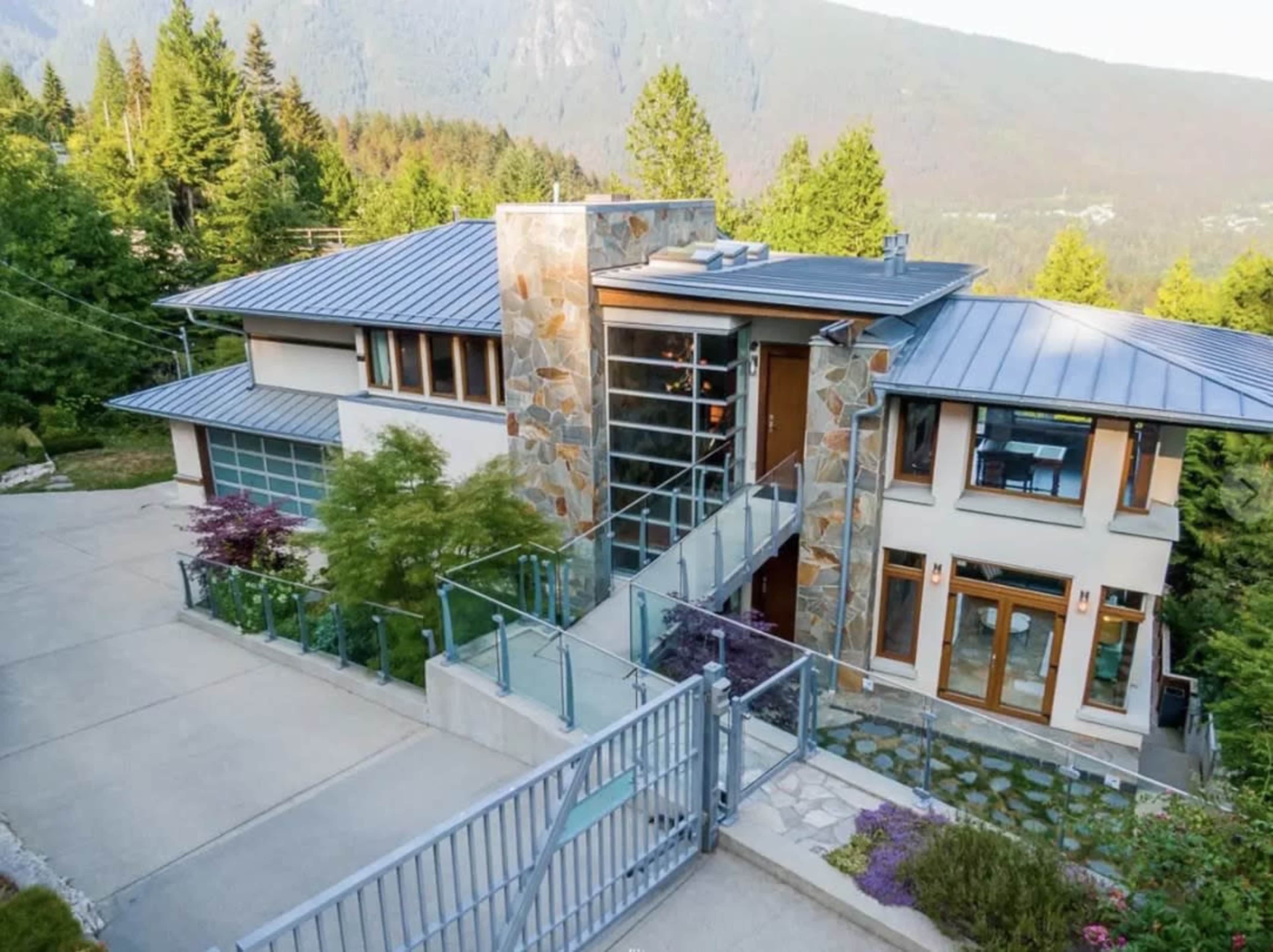 The image shows a modern two-story house with a stone and metal façade, surrounded by trees and mountainous terrain.