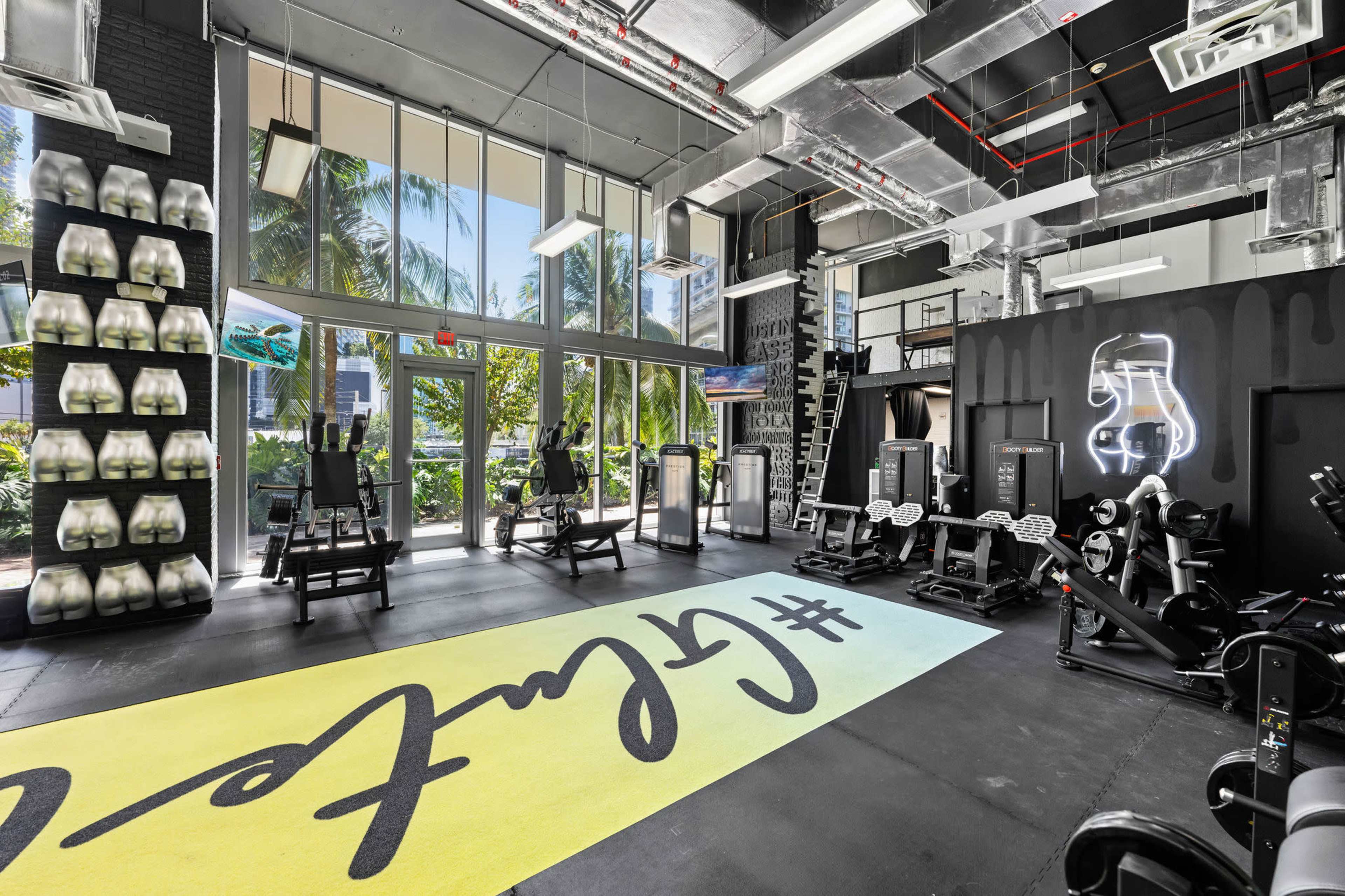A modern gym with various exercise equipment and large windows letting in natural light, surrounded by palm trees outside.