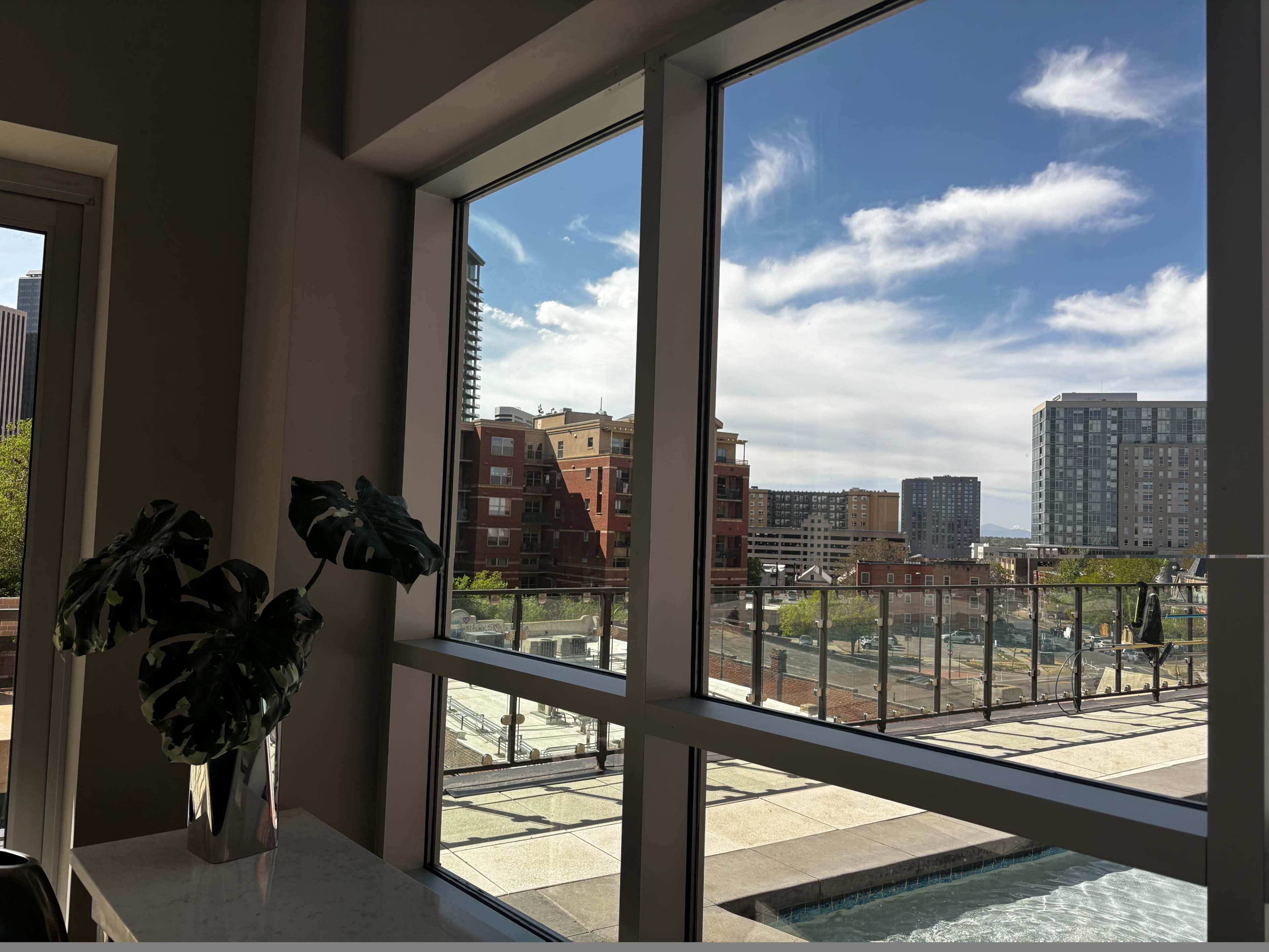 The scene shows a modern interior with large windows offering a view of an urban landscape, featuring buildings and a clear sky with clouds.