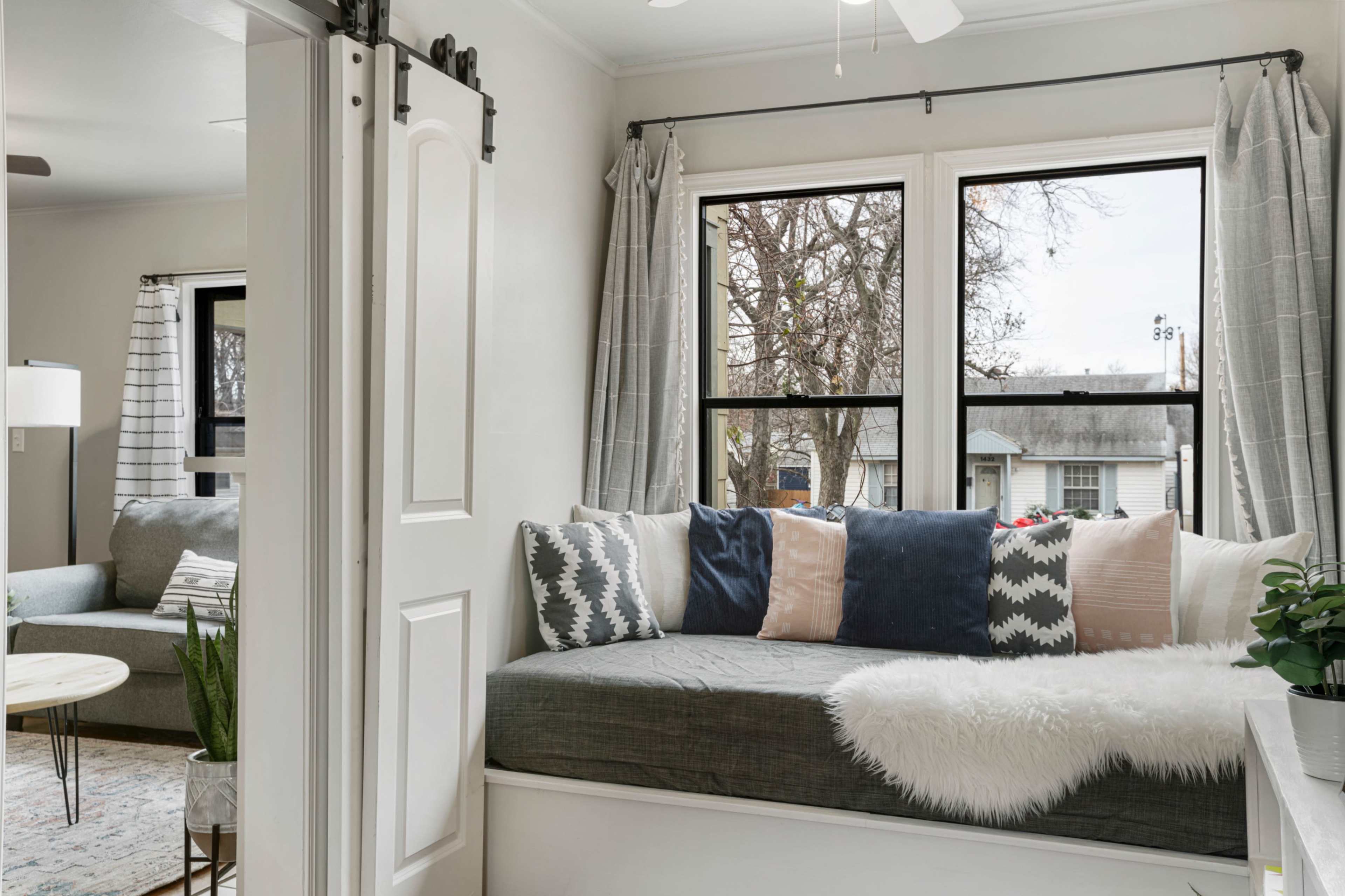 A cozy nook with a built-in bench is adorned with pillows and a faux fur throw, situated beside large windows that overlook an outdoor scene.