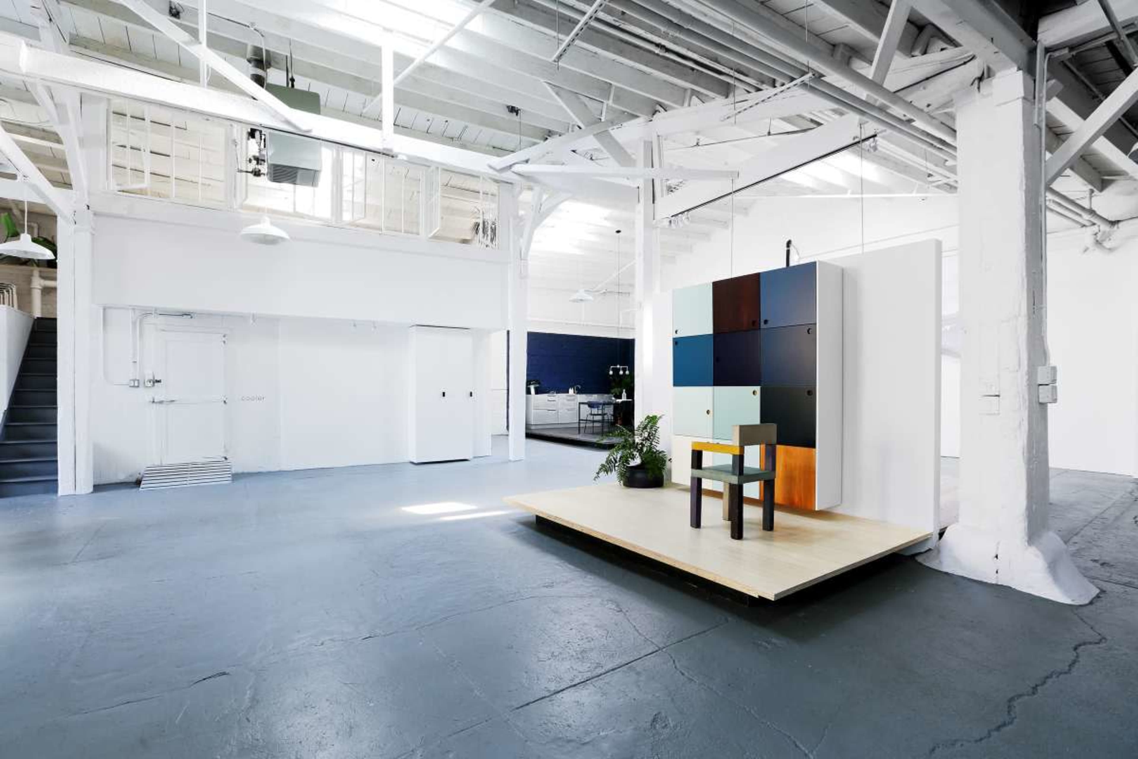 The image shows a spacious, industrial-style room with high ceilings, featuring a wooden platform with a chair and a color-blocked wall display, surrounded by white walls and large windows.