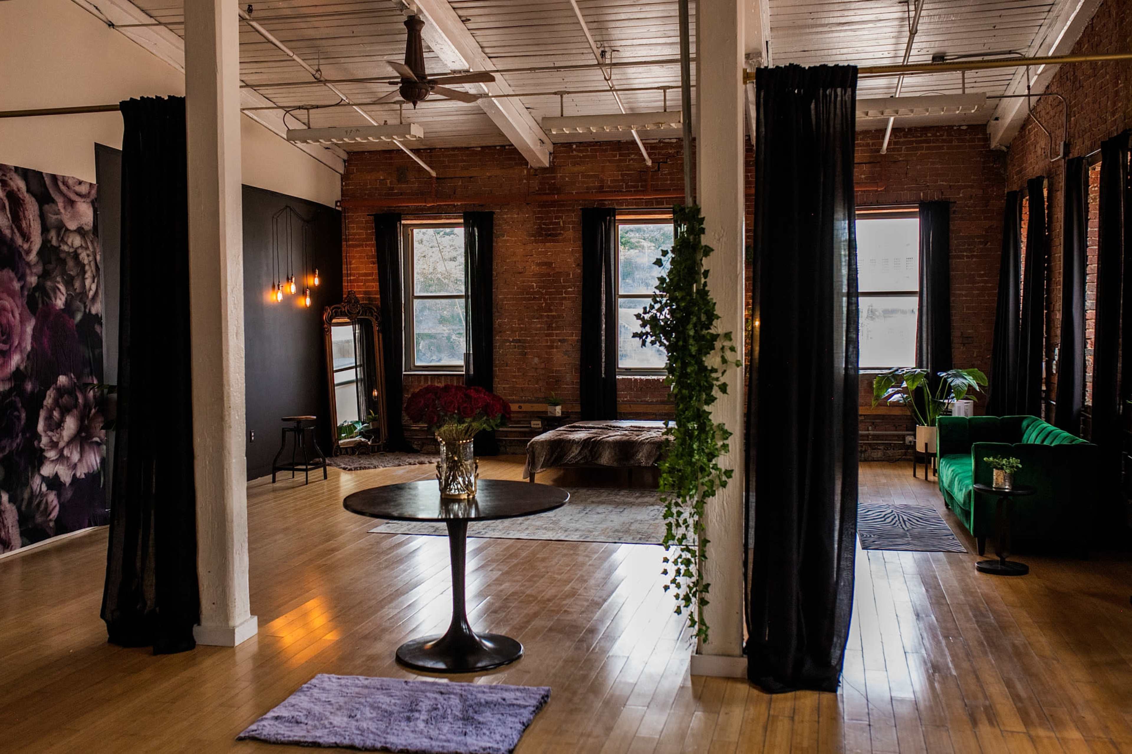 The image shows a spacious, modern interior with exposed brick walls, large windows, and a mix of greenery and stylish furniture.