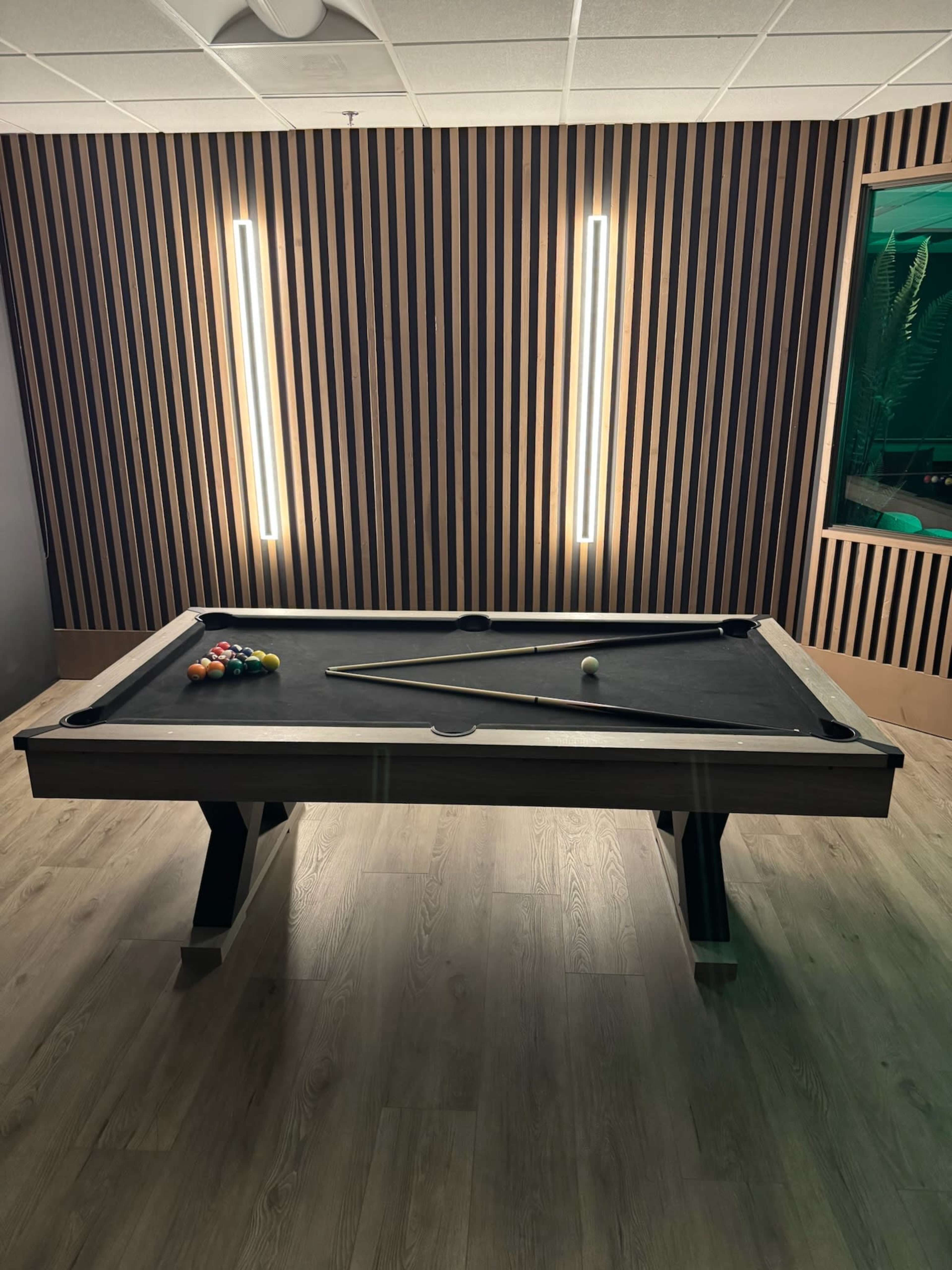 The image shows a billiard table with balls arranged on its surface, positioned in a room featuring wooden slat walls and bright overhead lights.