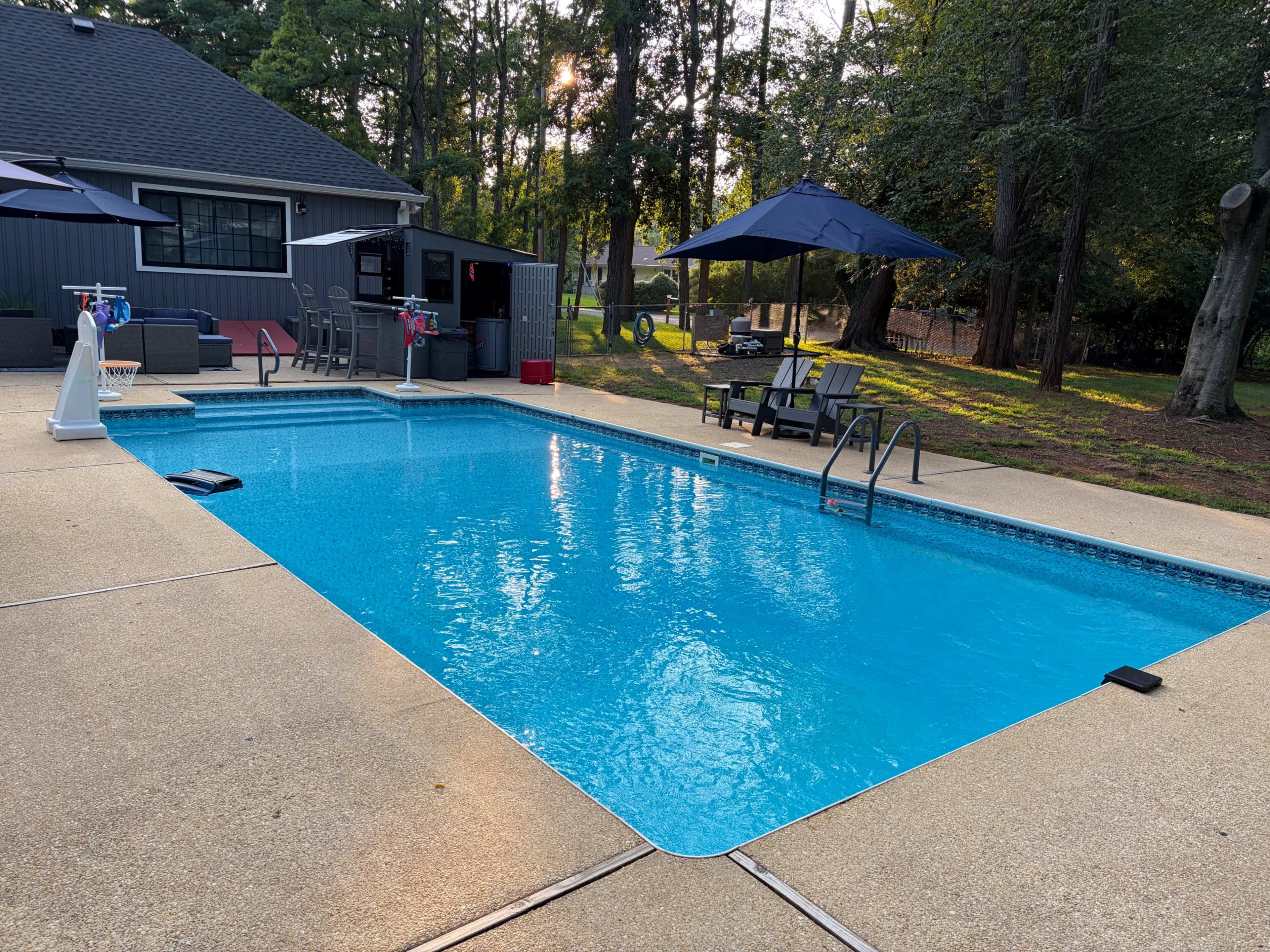 The image shows a rectangular swimming pool surrounded by concrete, with lounge chairs and umbrellas nearby, set in a wooded backyard area.
