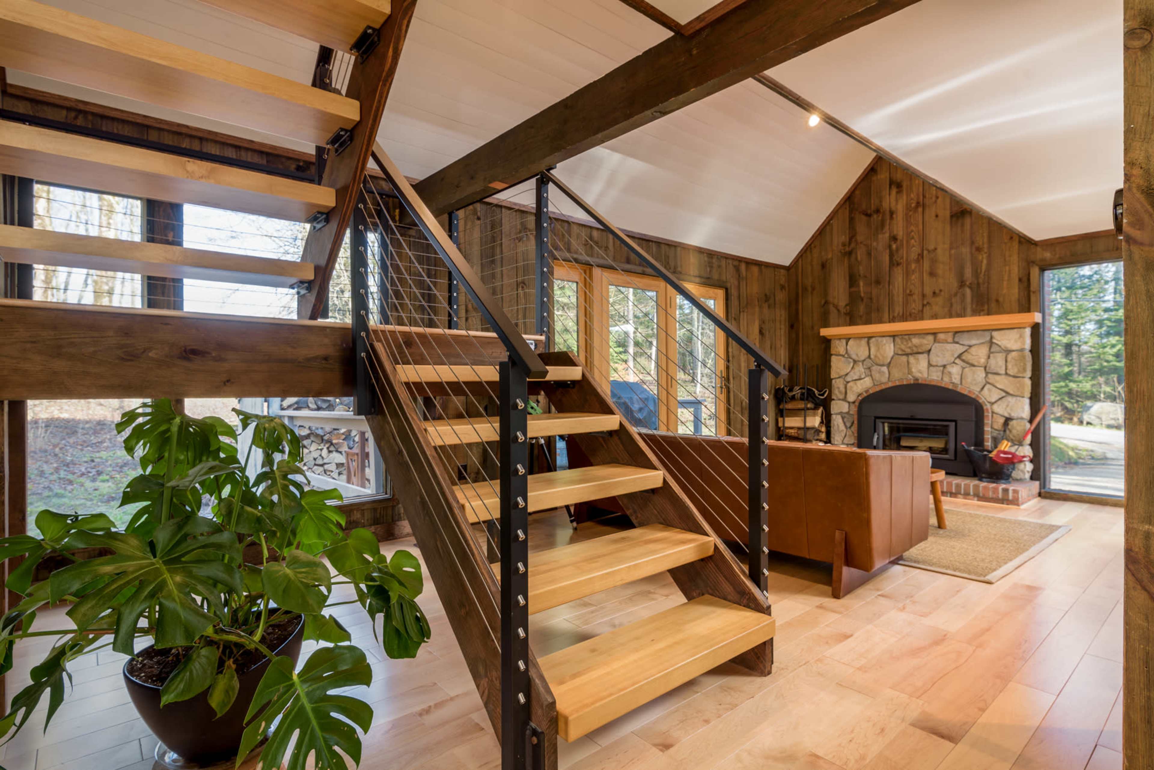 A modern interior with a wooden staircase, a stone fireplace, and large windows overlooking a wooded area.