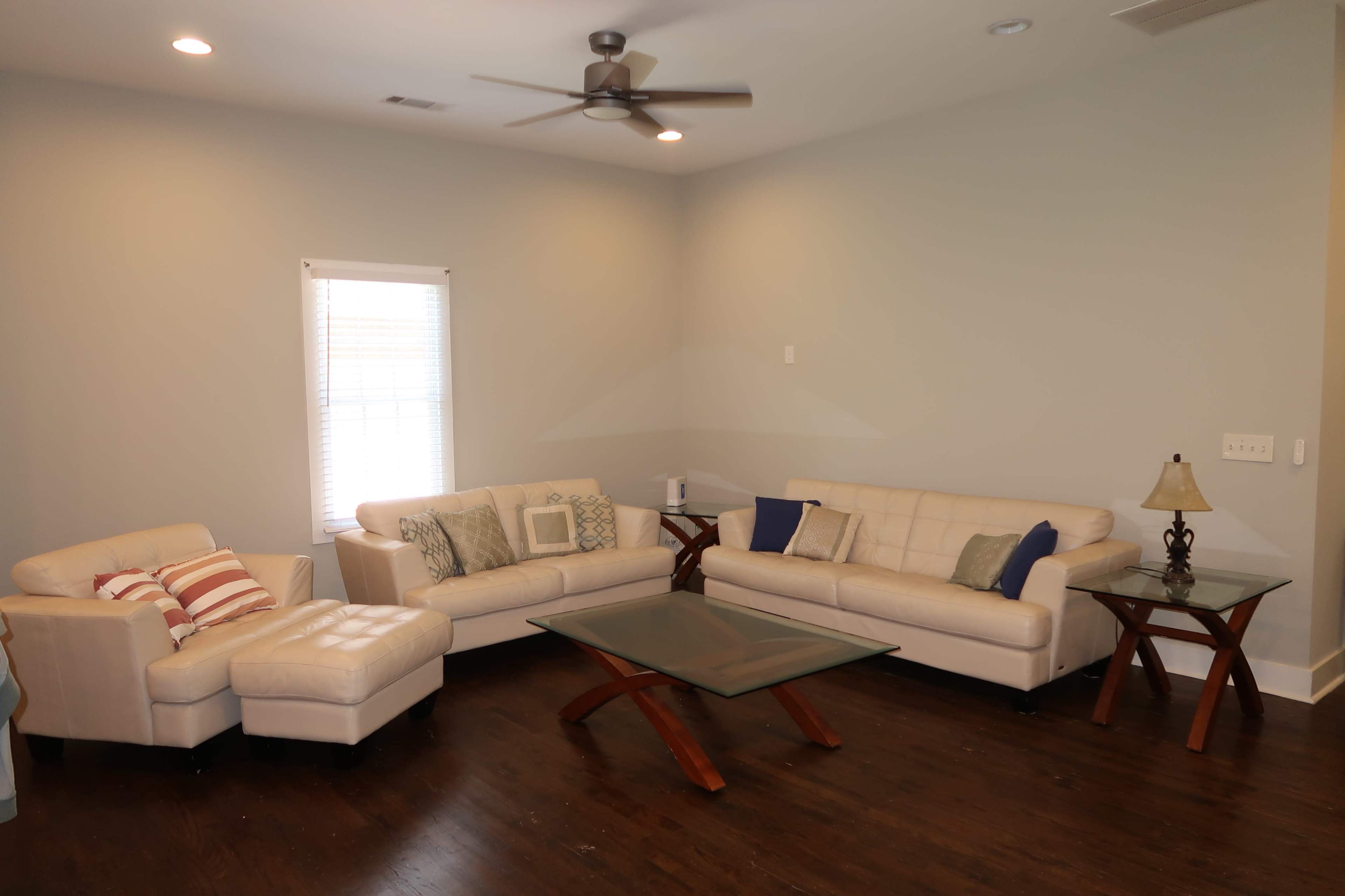 The image shows a living room with two light-colored leather sofas, a glass coffee table, a wooden side table with a lamp, and hardwood flooring.