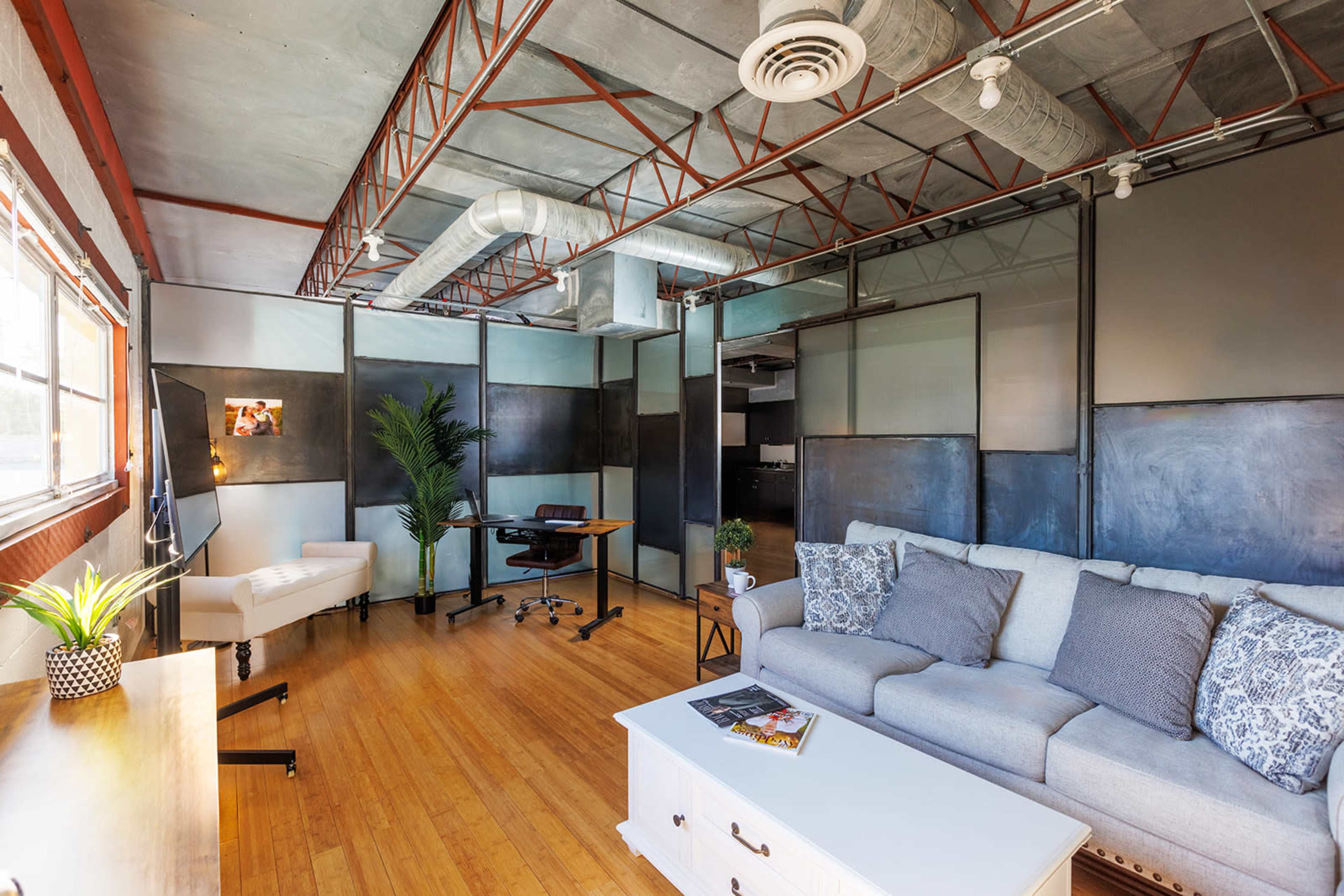 A modern office space featuring a grey couch, a wooden coffee table, a desk, and a large window with natural light, surrounded by industrial-style decor.