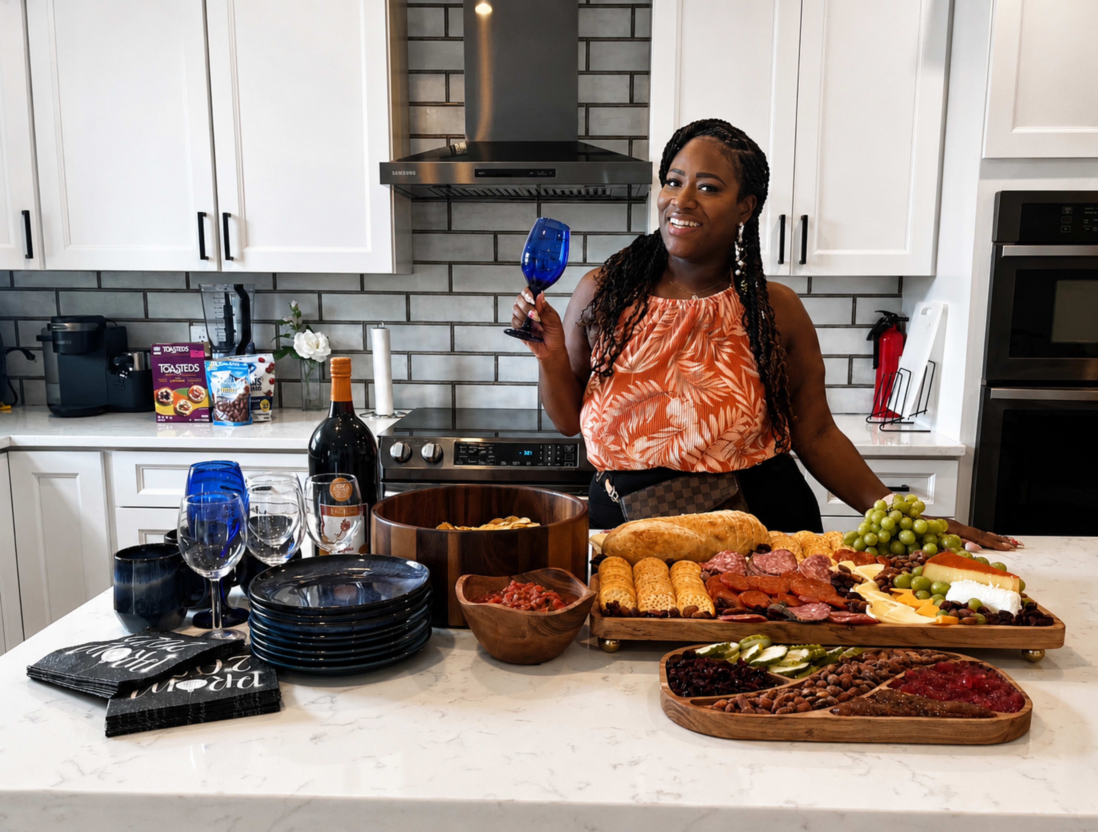 A woman stands in a modern kitchen holding a blue glass, surrounded by a table filled with various foods and drinks, including cheeses, meats, fruits, and bread.