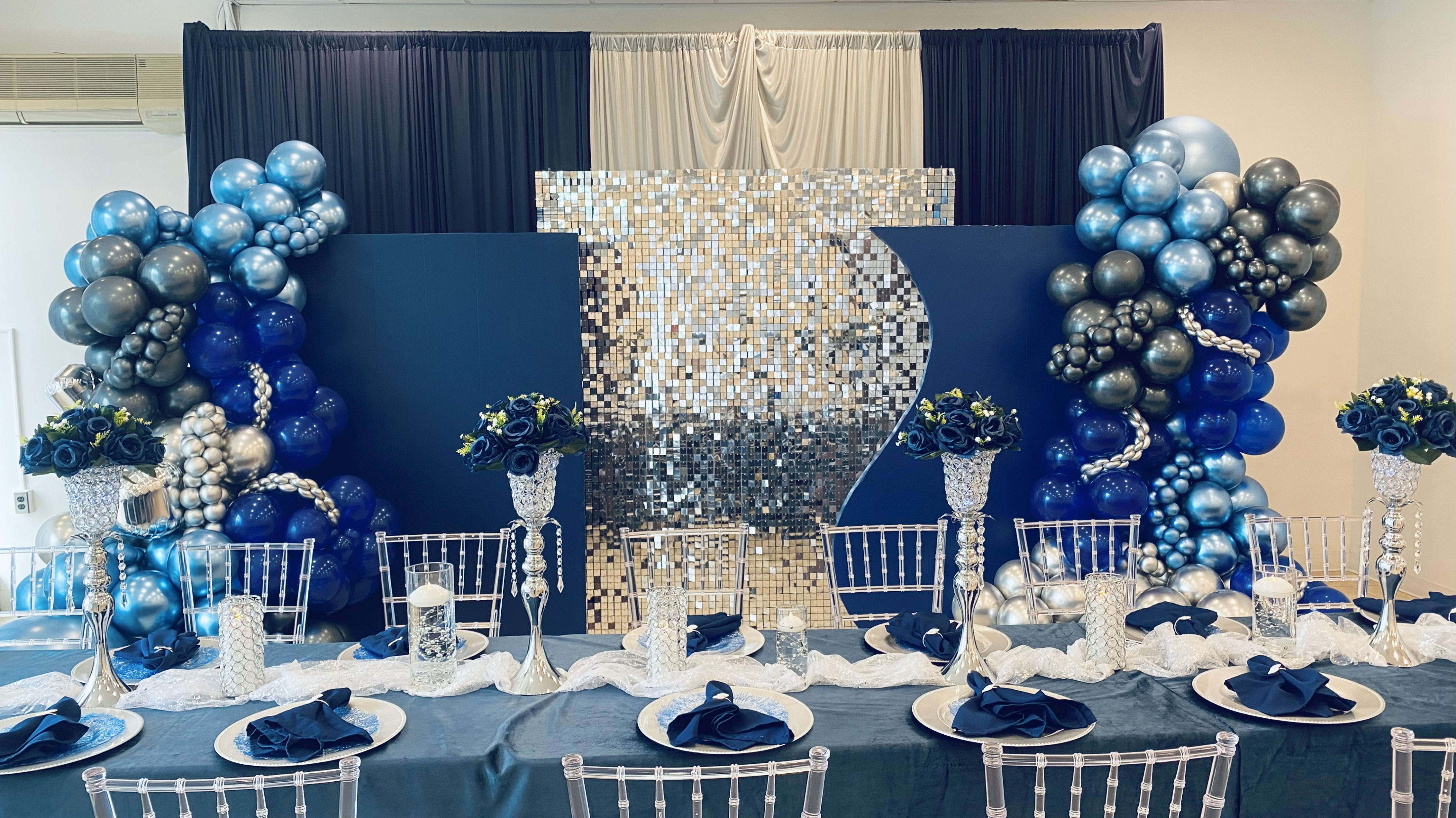 The image shows a decorated event space featuring a mirrored backdrop surrounded by blue and silver balloons, with an elegantly set table in front.