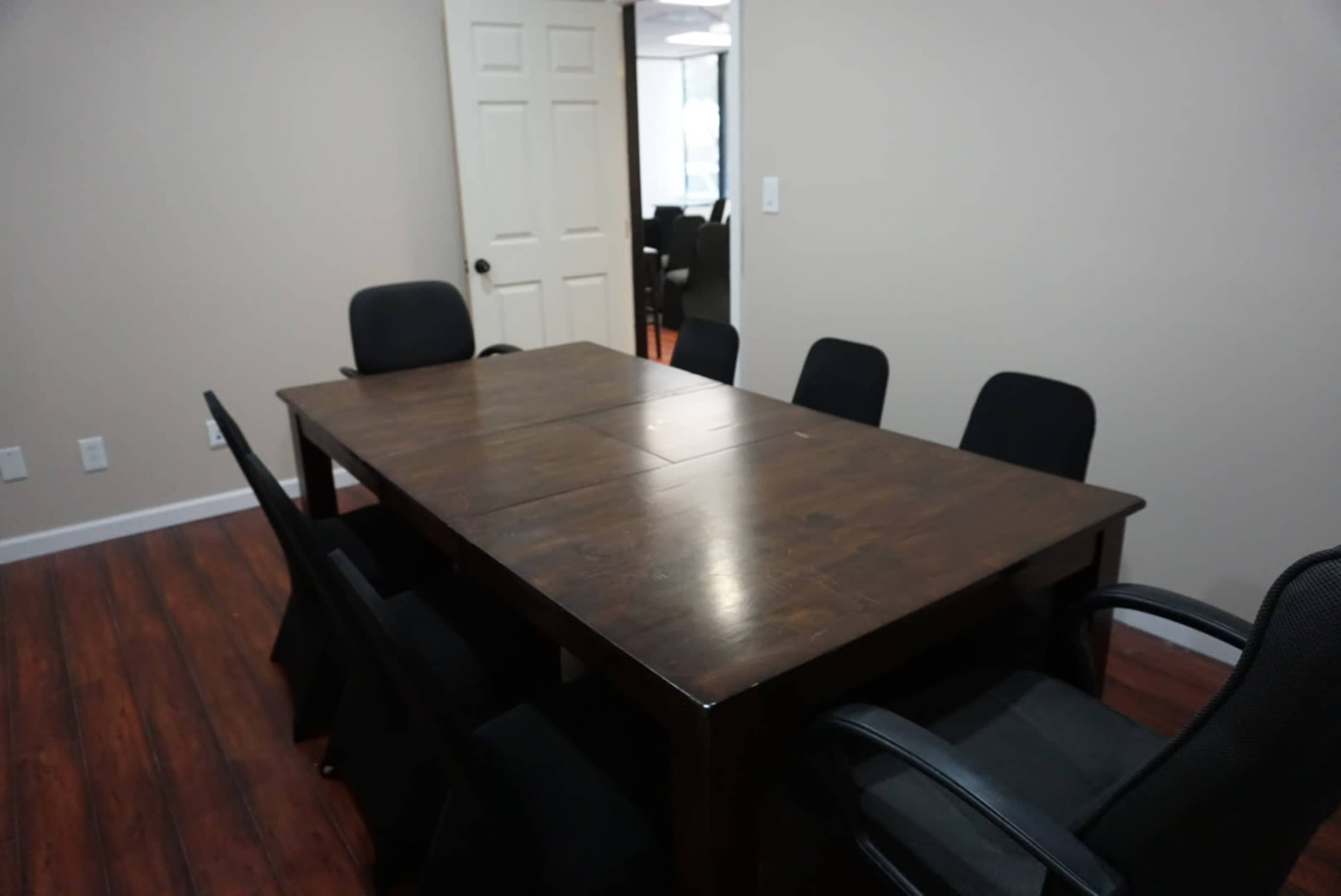 A wooden conference table is surrounded by six black chairs in a sparsely furnished room.
