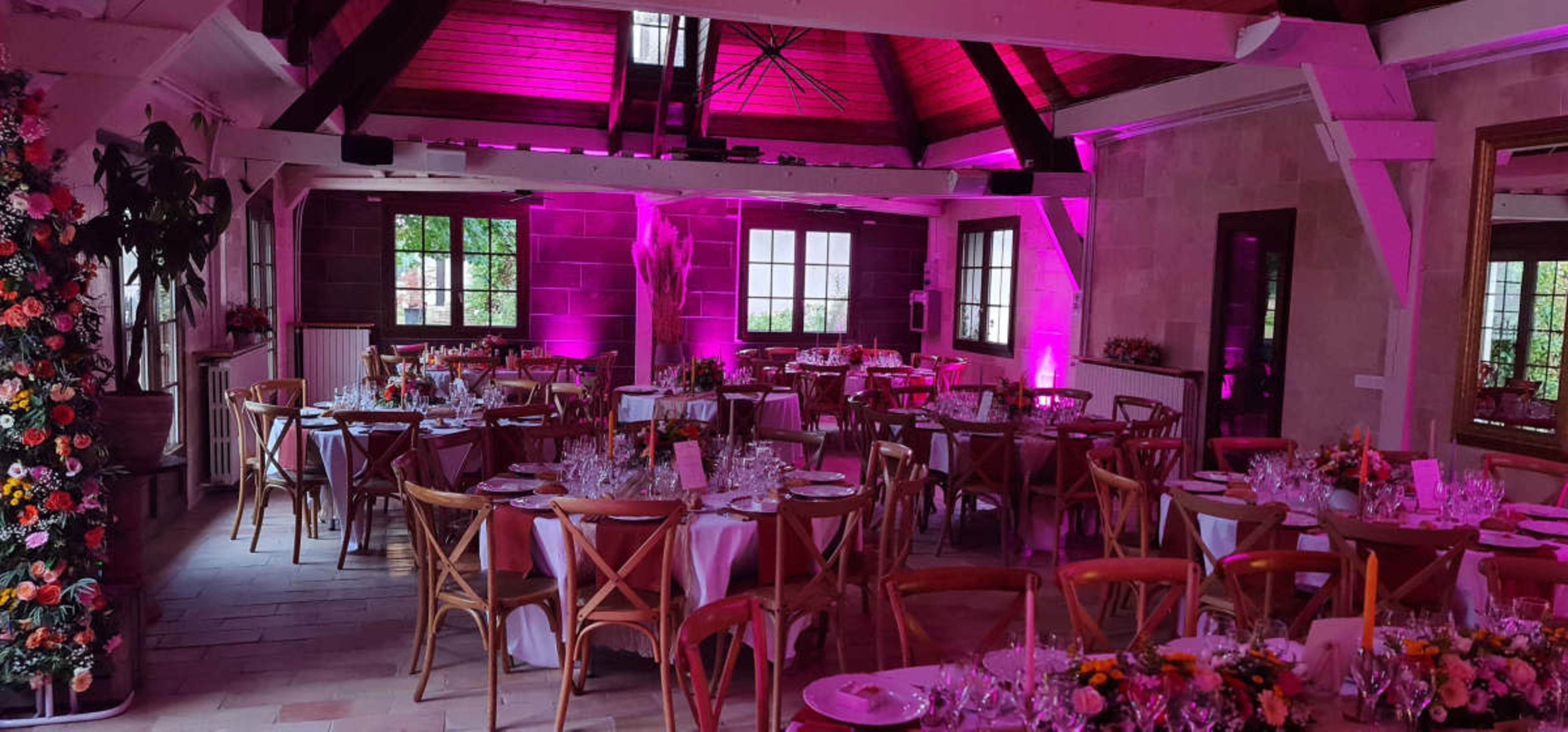 The image shows a dining area with tables set for an event, featuring floral centerpieces and pink lighting.
