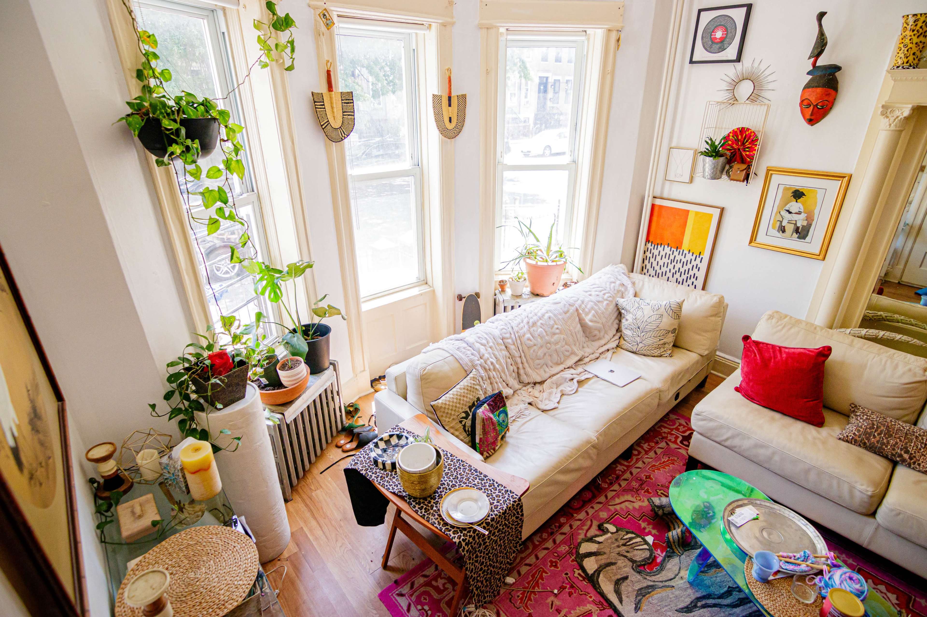 A well-lit living room features a white sofa, colorful decor, houseplants, and a mix of artwork on the walls.