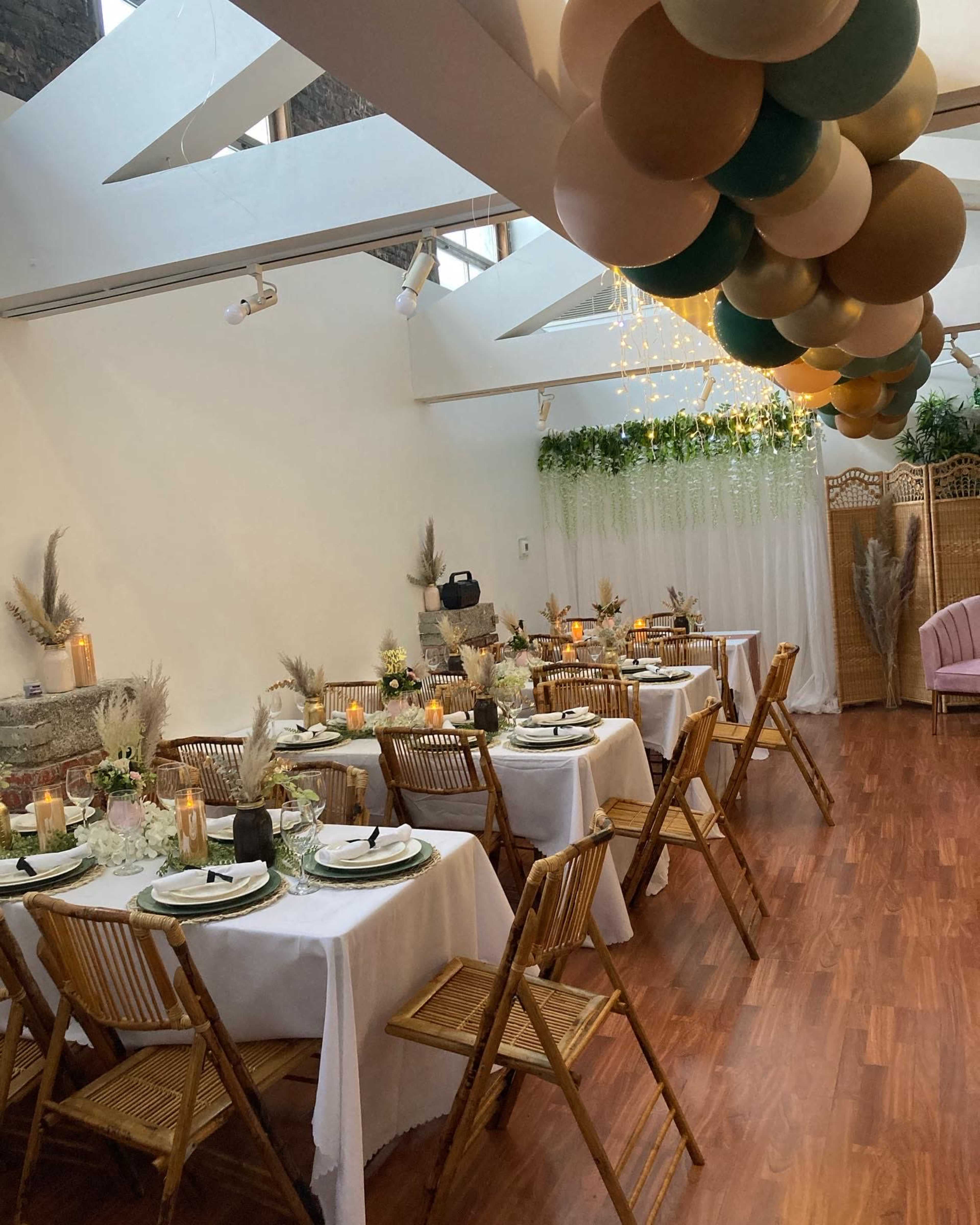 The image shows a decorated event space with several tables set for dining, featuring floral centerpieces, candles, and a balloon arrangement hanging above.