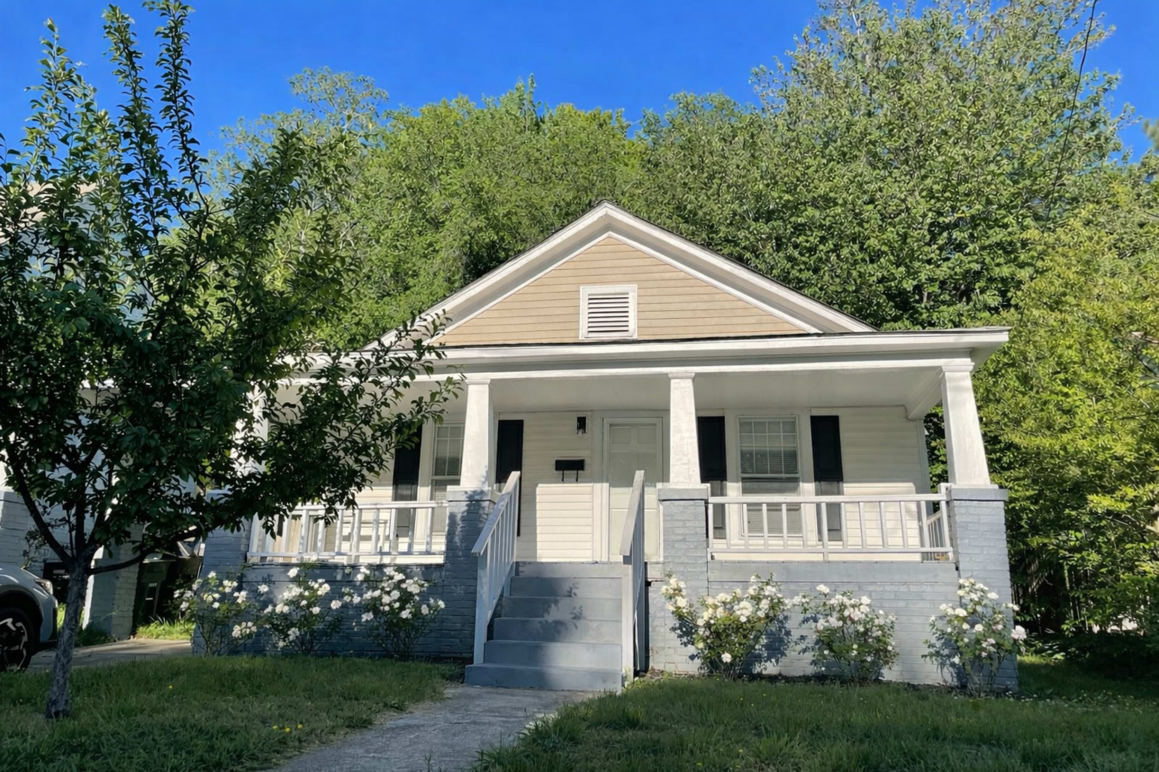 The image shows a single-story house with a front porch, surrounded by greenery and blooming rose bushes.