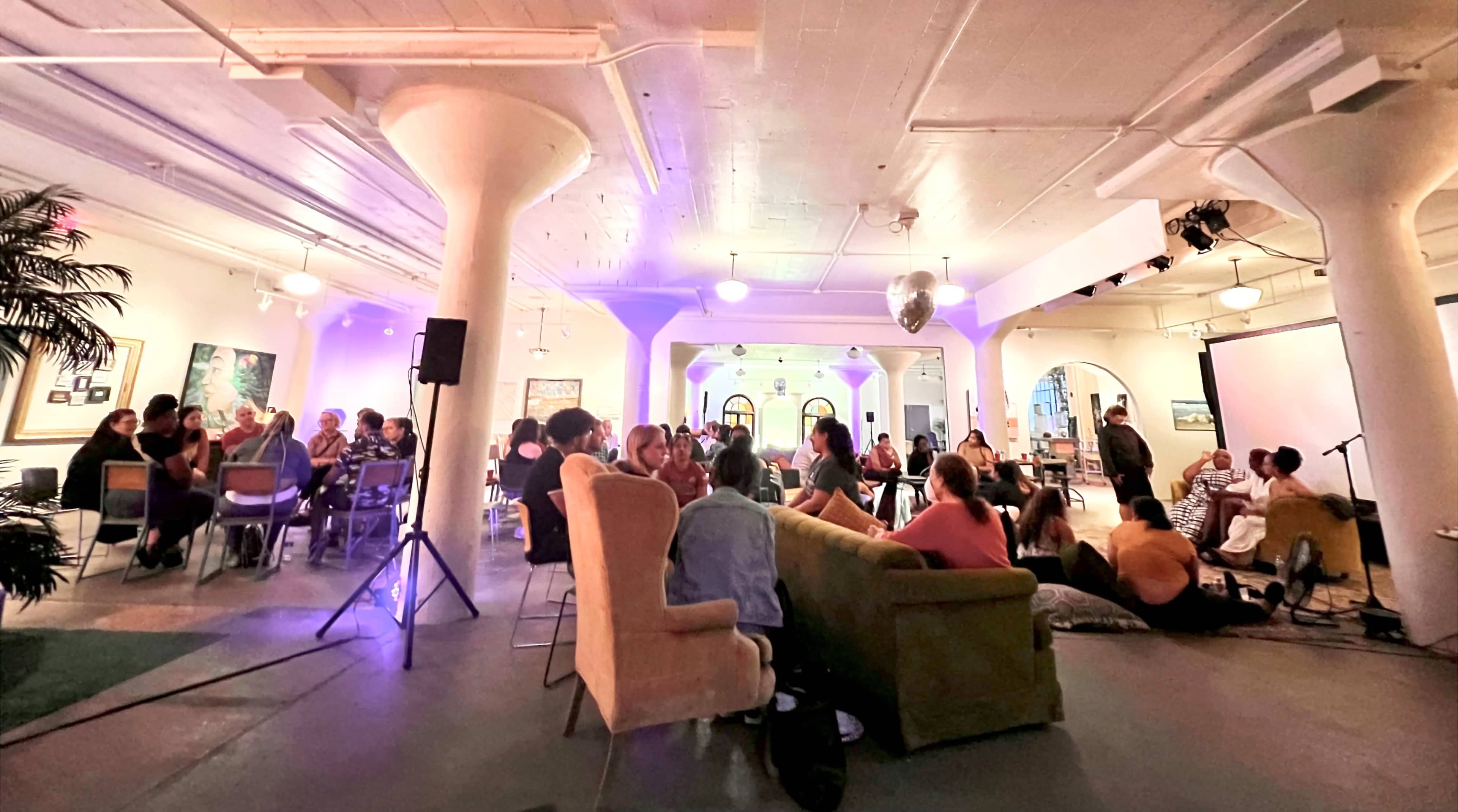 A large indoor space filled with people gathered around seating areas, socializing under soft purple lighting.