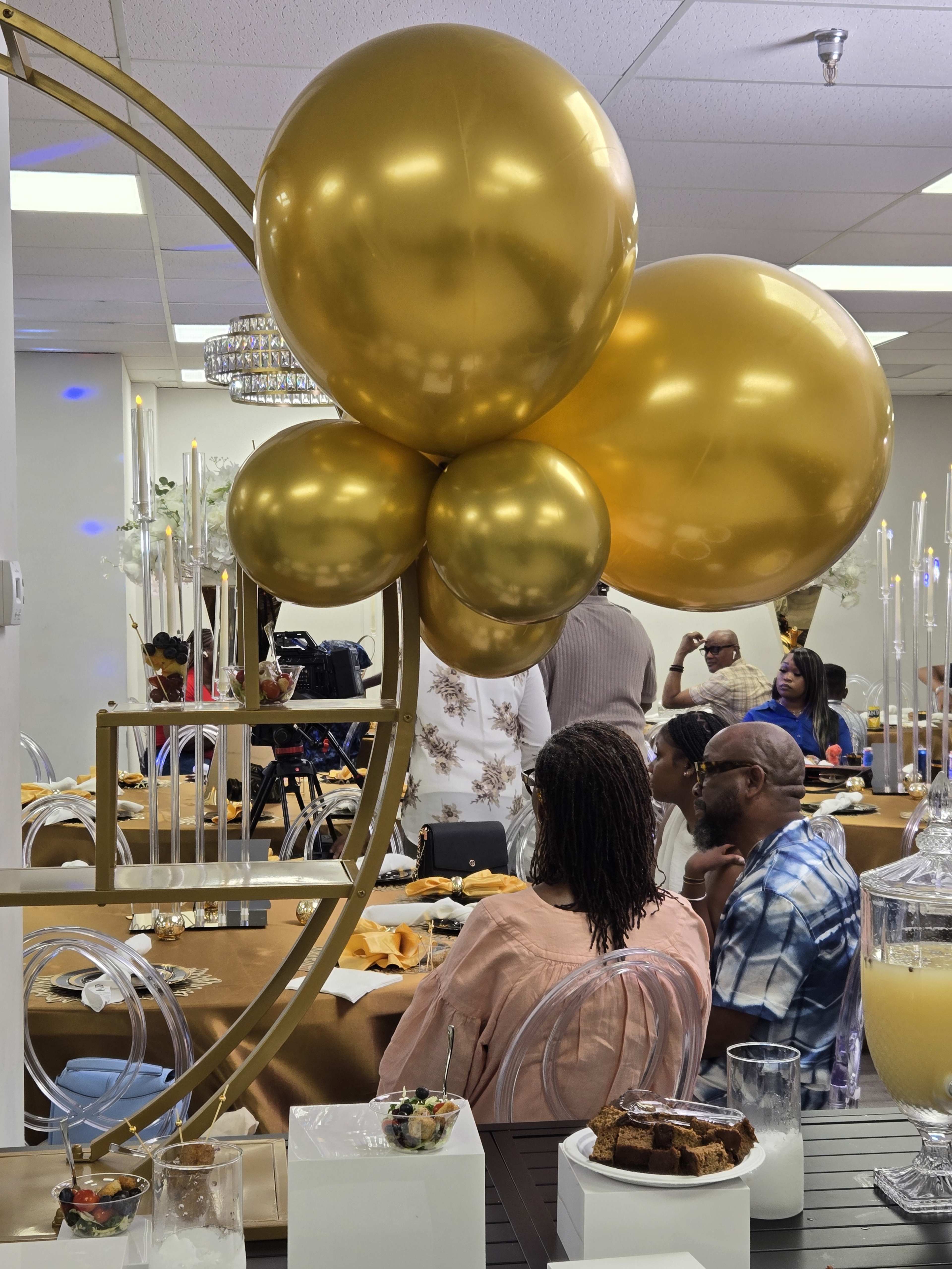 A cluster of gold balloons is positioned in the foreground while guests sit at decorated tables in a festive setting.