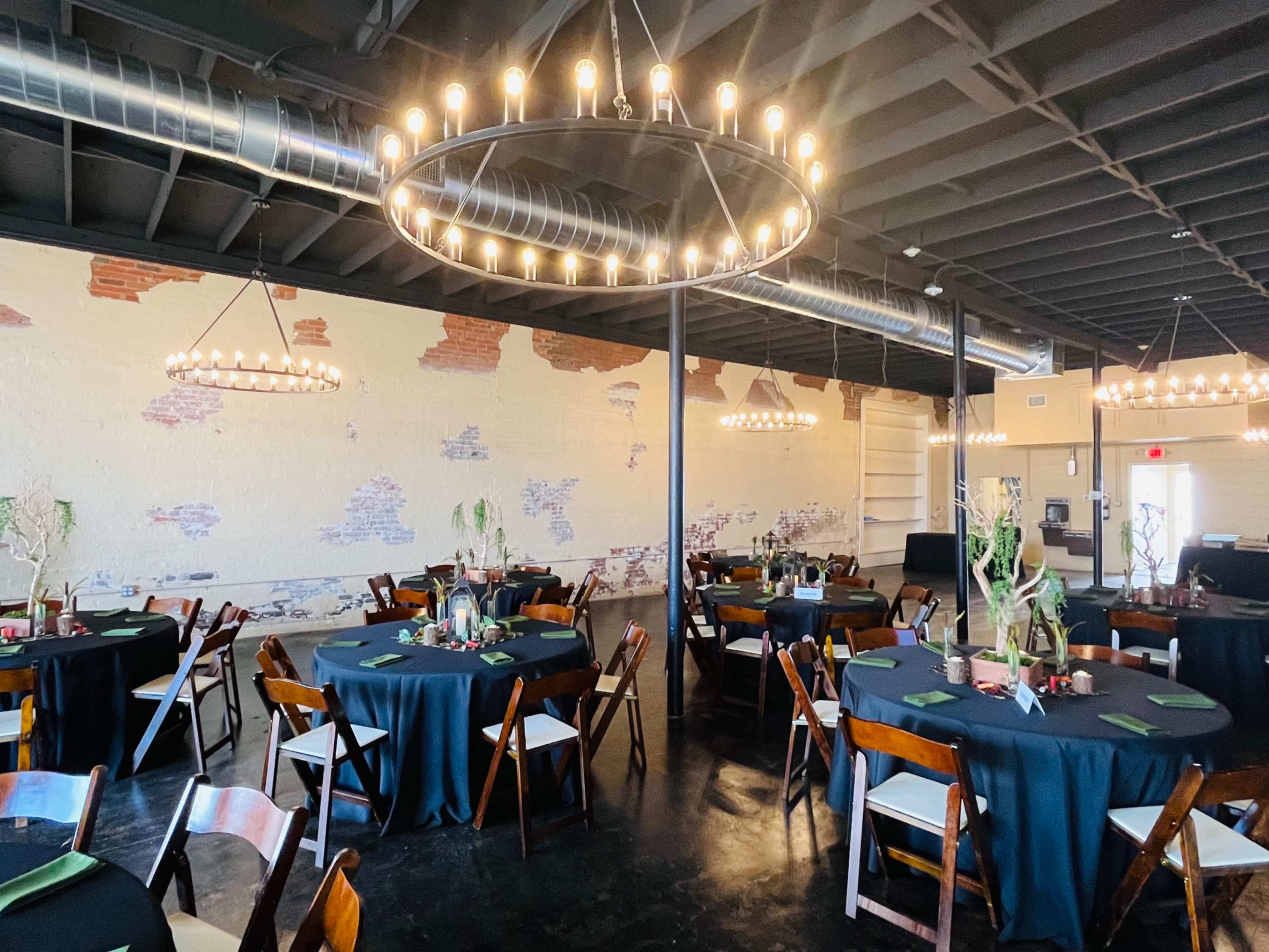 The image shows a spacious event venue with round tables covered in black tablecloths, each set with green napkins, and illuminated by large chandeliers hanging from the ceiling.