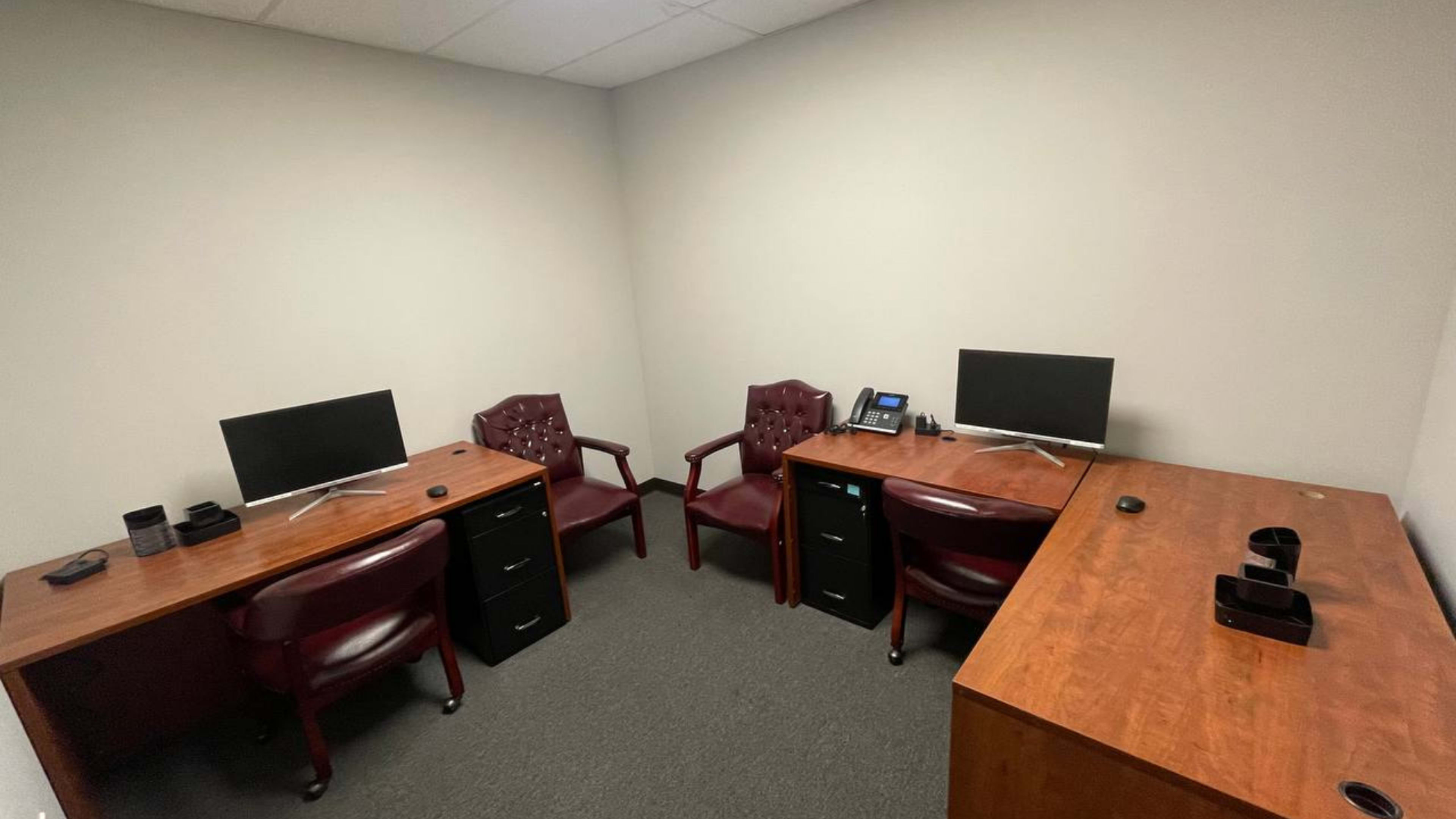 The image shows a small office space with two desks, each equipped with a computer monitor and a phone, and two burgundy chairs adjacent to the desks.