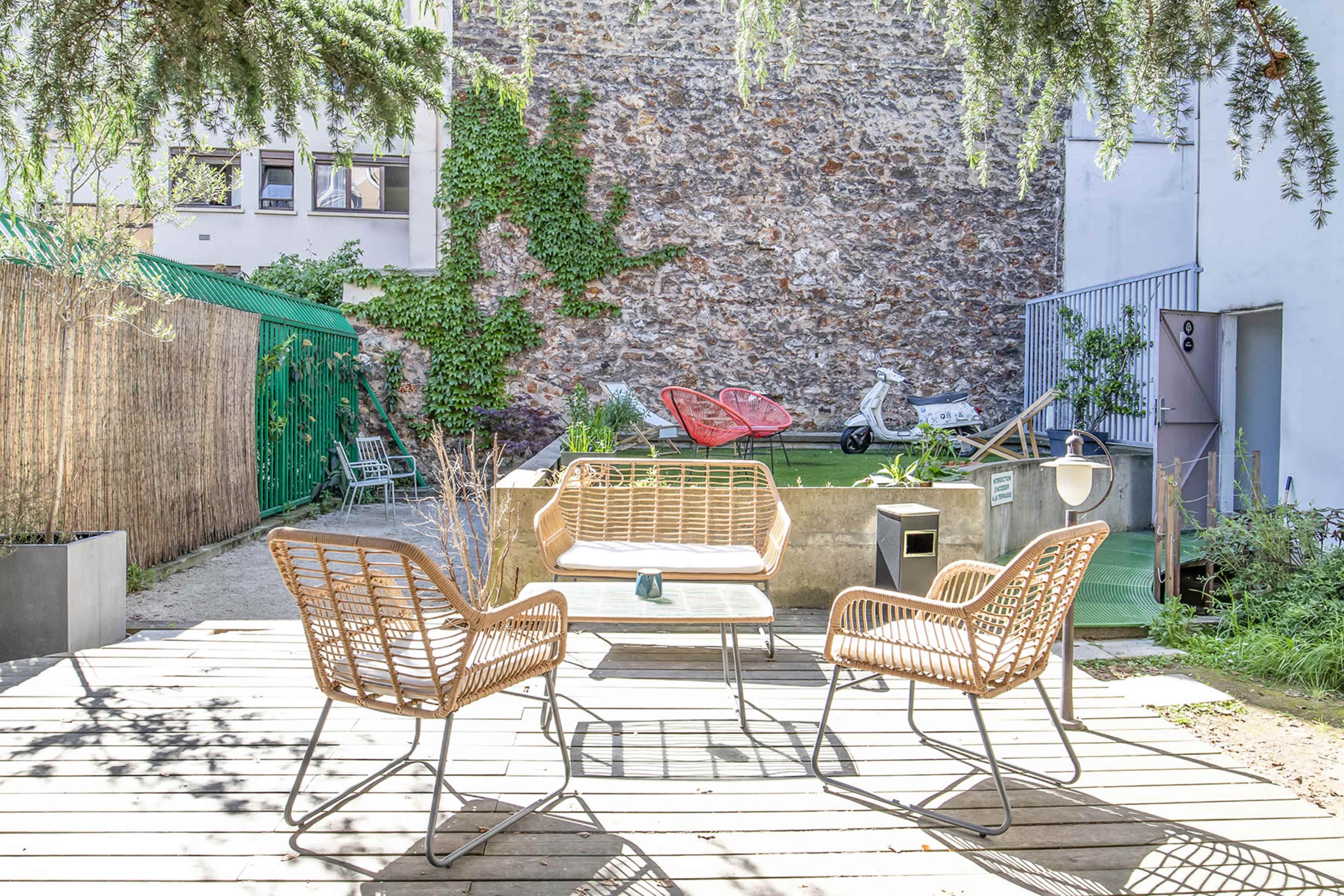 A courtyard with wooden decking, featuring a table and four woven chairs, surrounded by a stone wall, greenery, and a few decorative elements.
