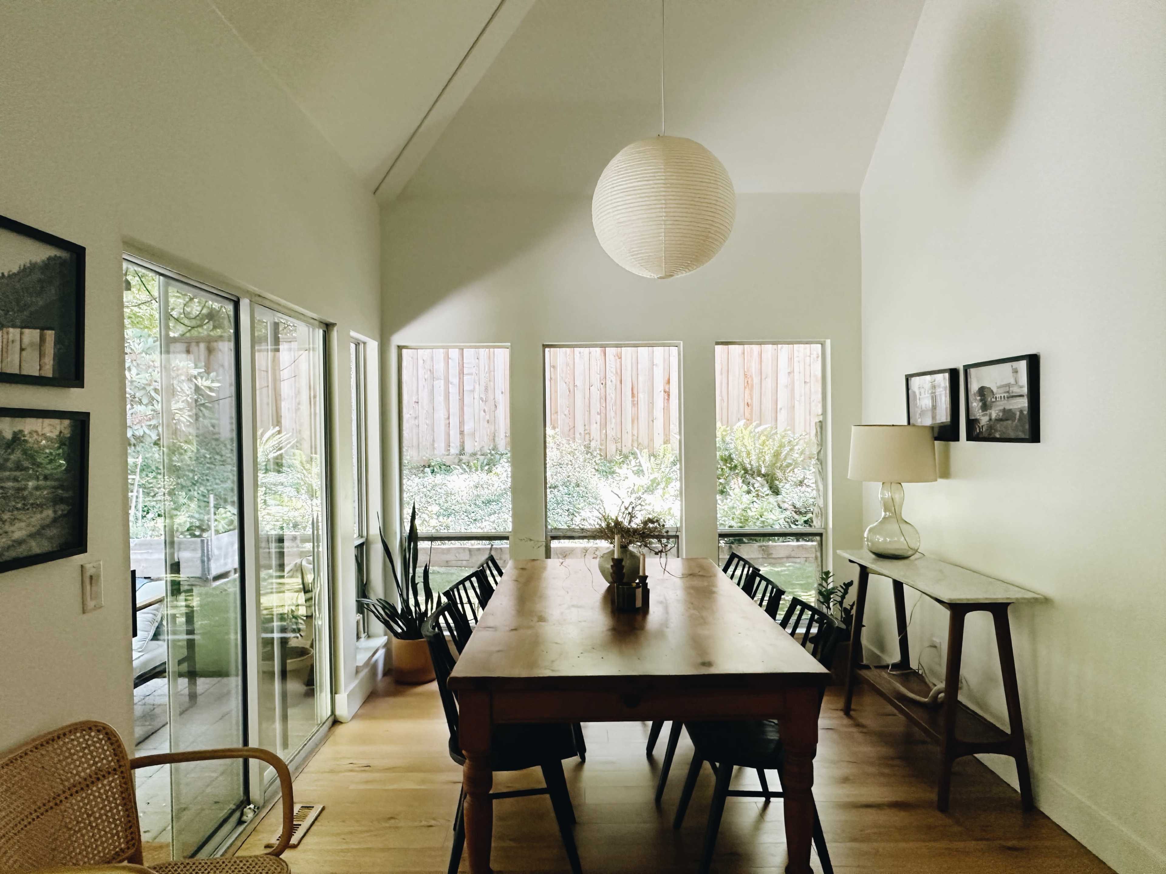 A dining area features a wooden table surrounded by chairs, with large windows providing views of a lush outdoor space.
