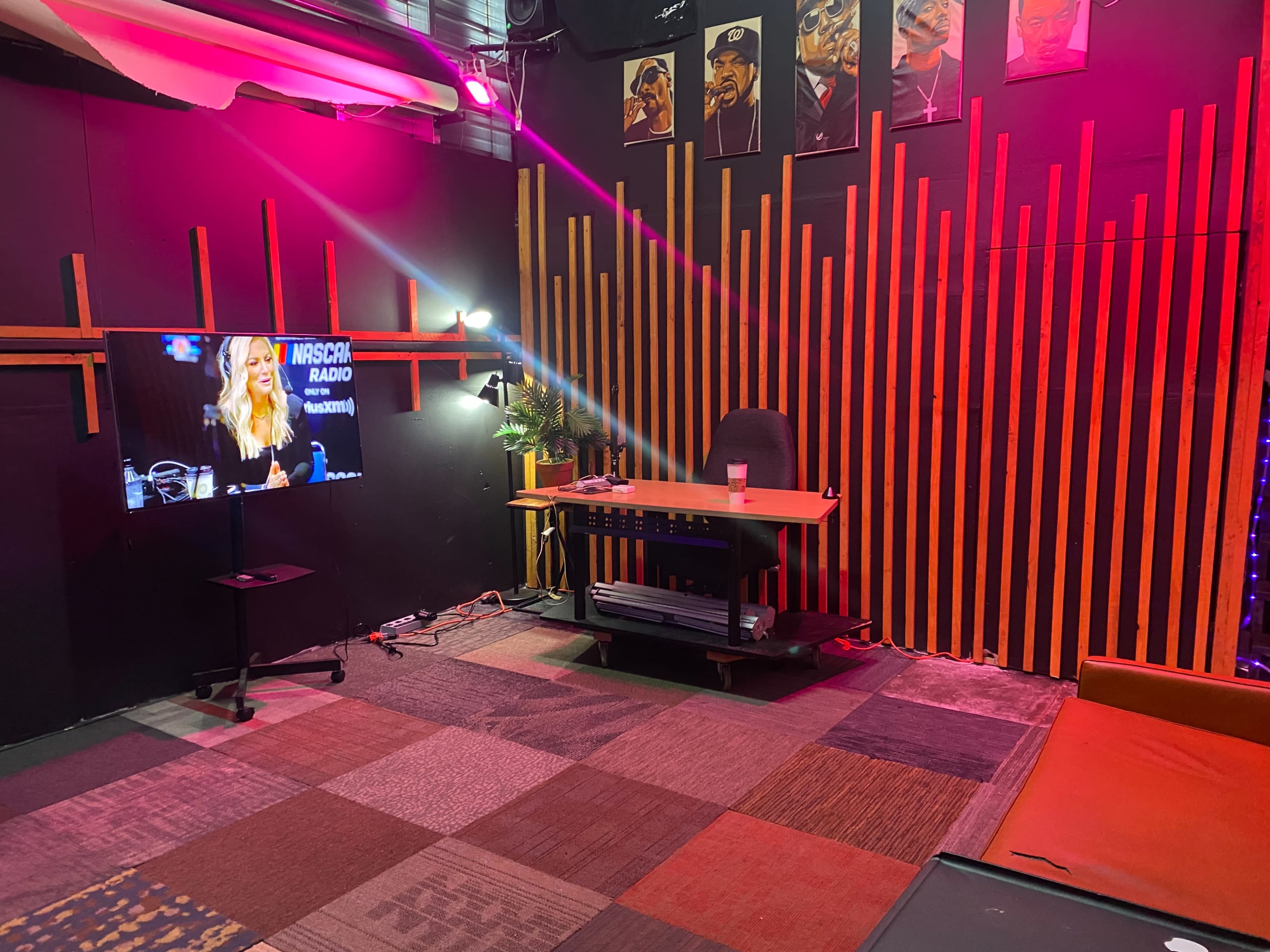 The image shows a studio or room designed for audio or video productions, featuring a wooden desk, a television displaying a program, and a backdrop of colorful vertical wooden strips.