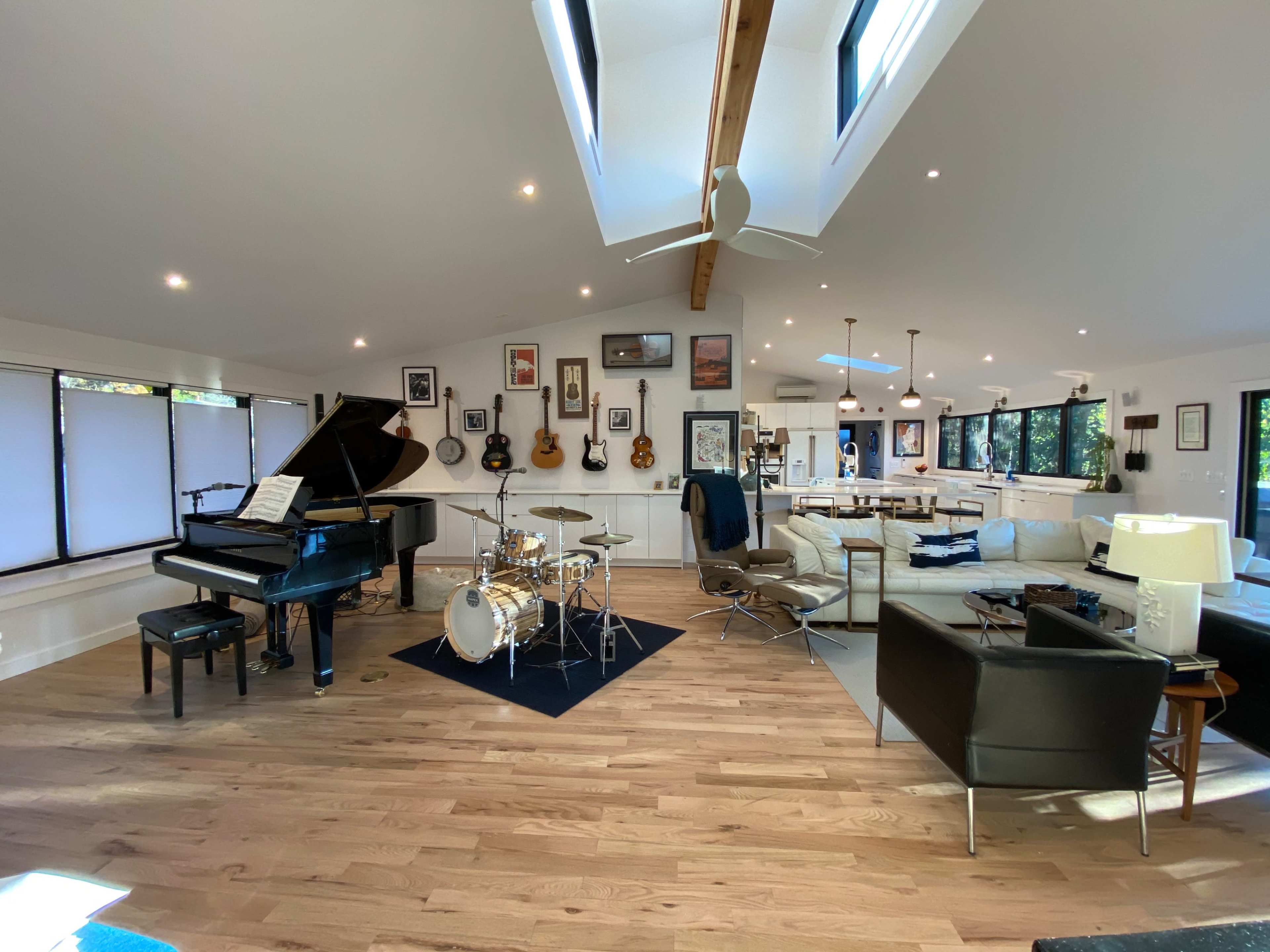 A spacious, well-lit room featuring a grand piano, a drum set, several guitars on the walls, and a mix of seating areas.