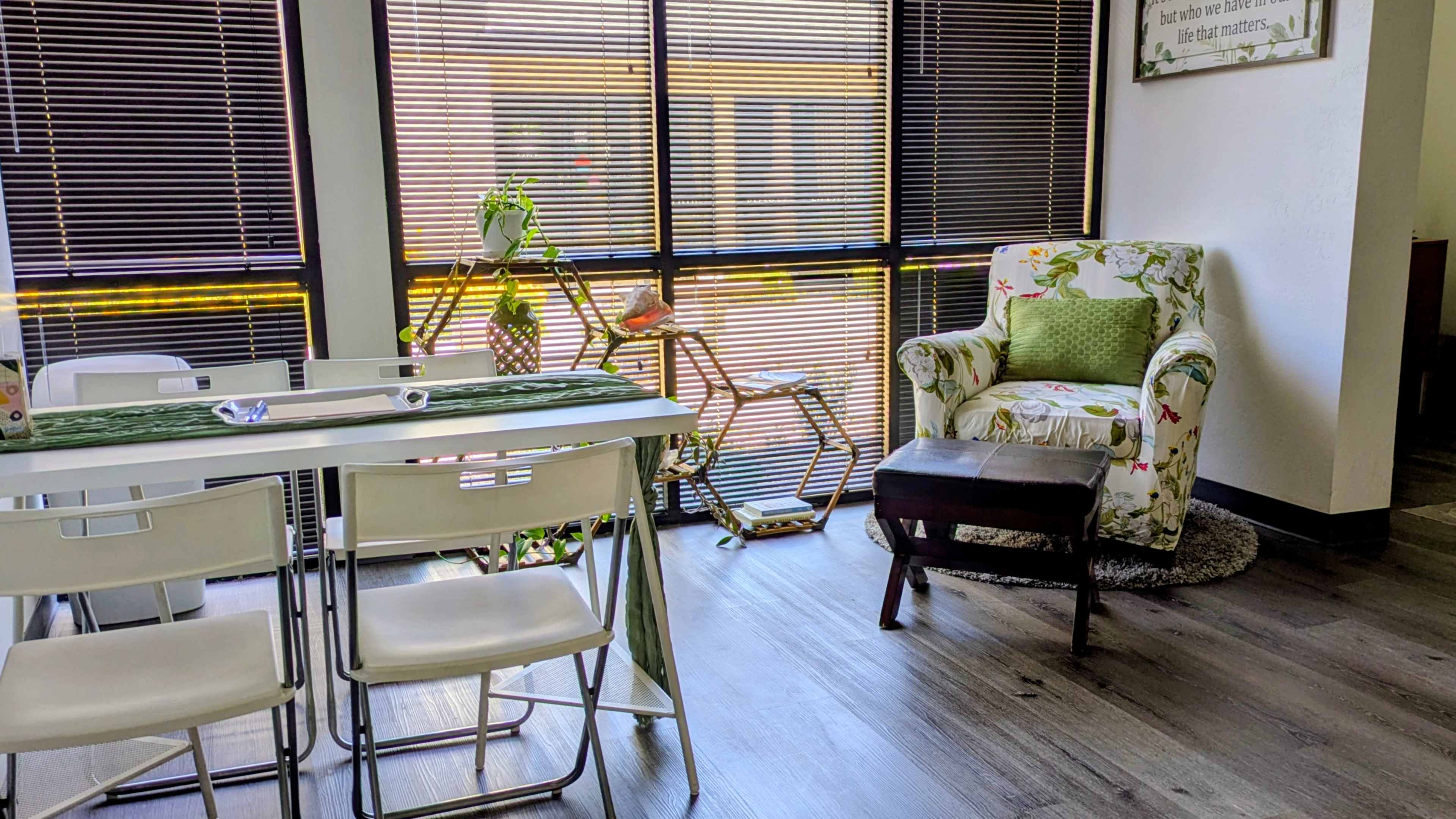 The image shows a well-lit room featuring a dining table with chairs, a patterned armchair, and a small footrest, all arranged near large windows with blinds.